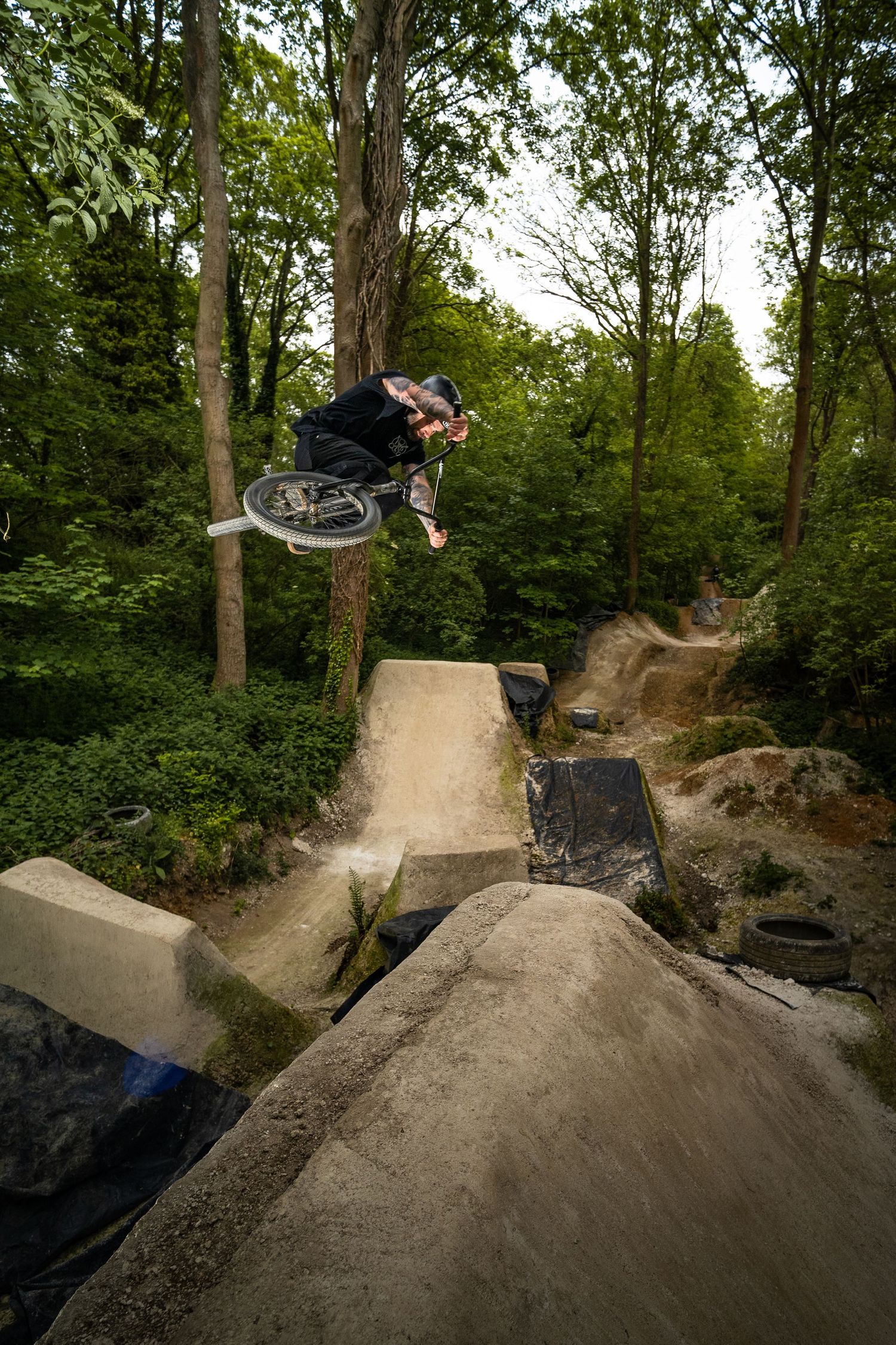 BMX rider performing an aerial trick over concrete jumps at an outdoor dirt bike park surrounded by trees.