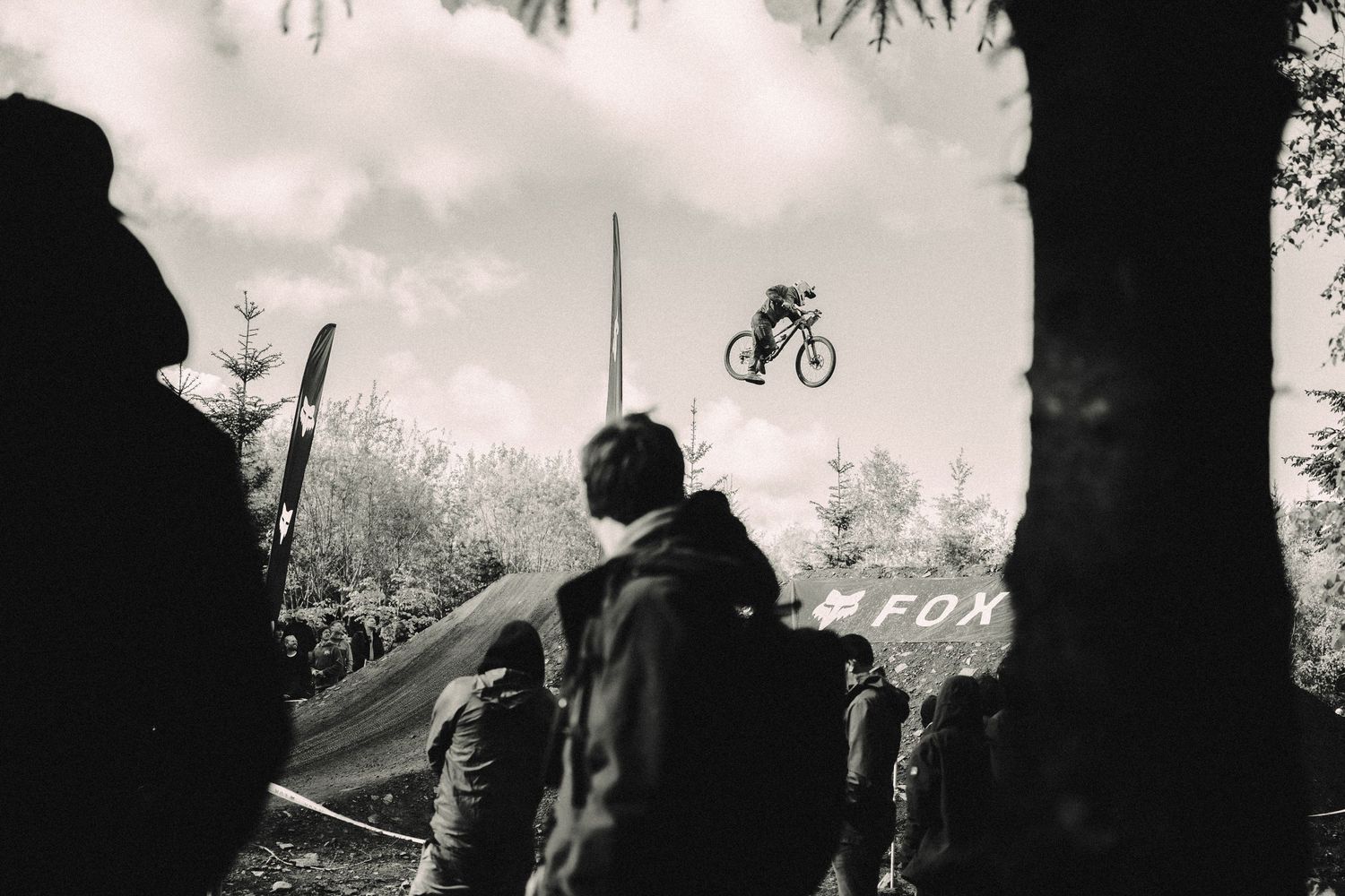 Black and white photo of a mountain biker performing a jump at a Fox-sponsored bike park event with spectators watching.