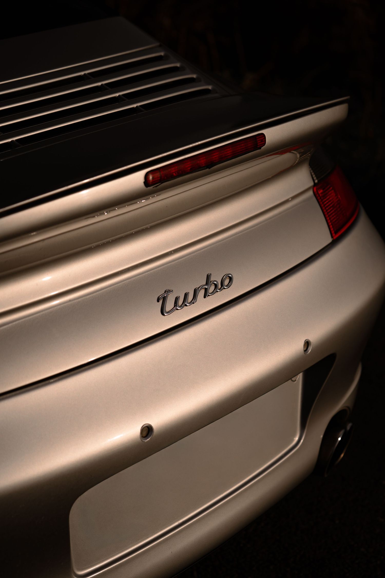 Close-up of a silver Porsche 911 Turbo rear badge and spoiler in dramatic low-key lighting.