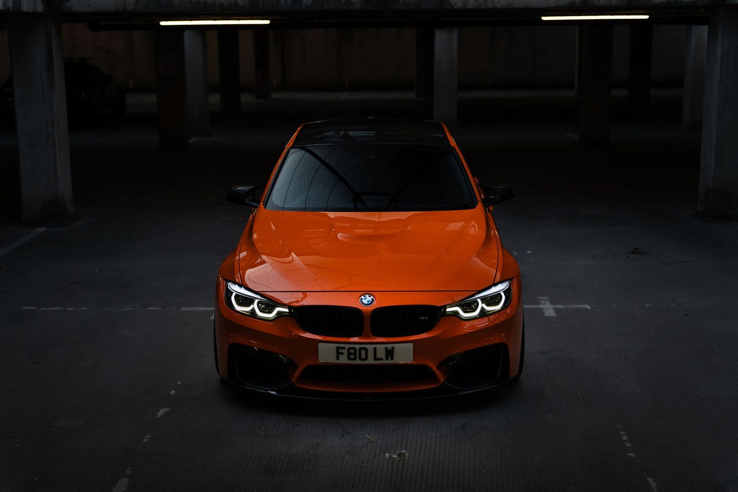 Vibrant orange BMW M4 parked in a dark garage, showcasing its iconic kidney grille and angel eye headlights.