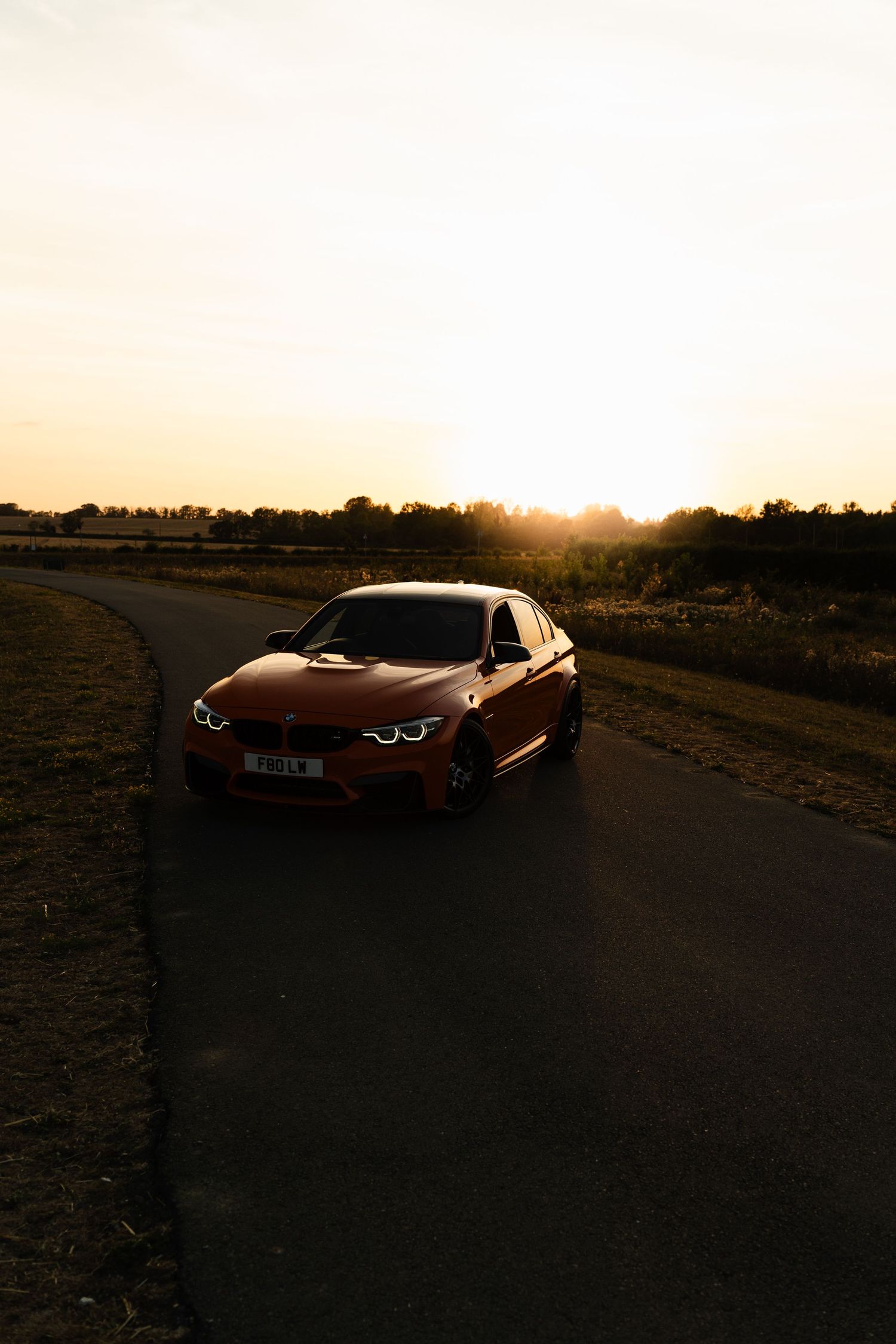 Red BMW M3 sedan parked on a winding road at golden sunset with warm light illuminating the landscape.
