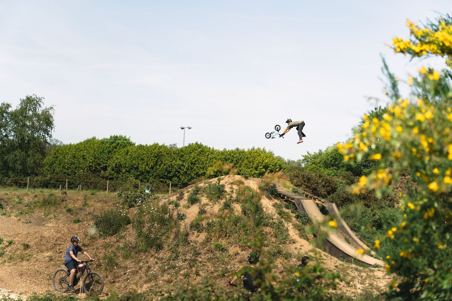 BMX rider performs aerial trick over dirt jumps while another cyclist watches at an outdoor bike park.