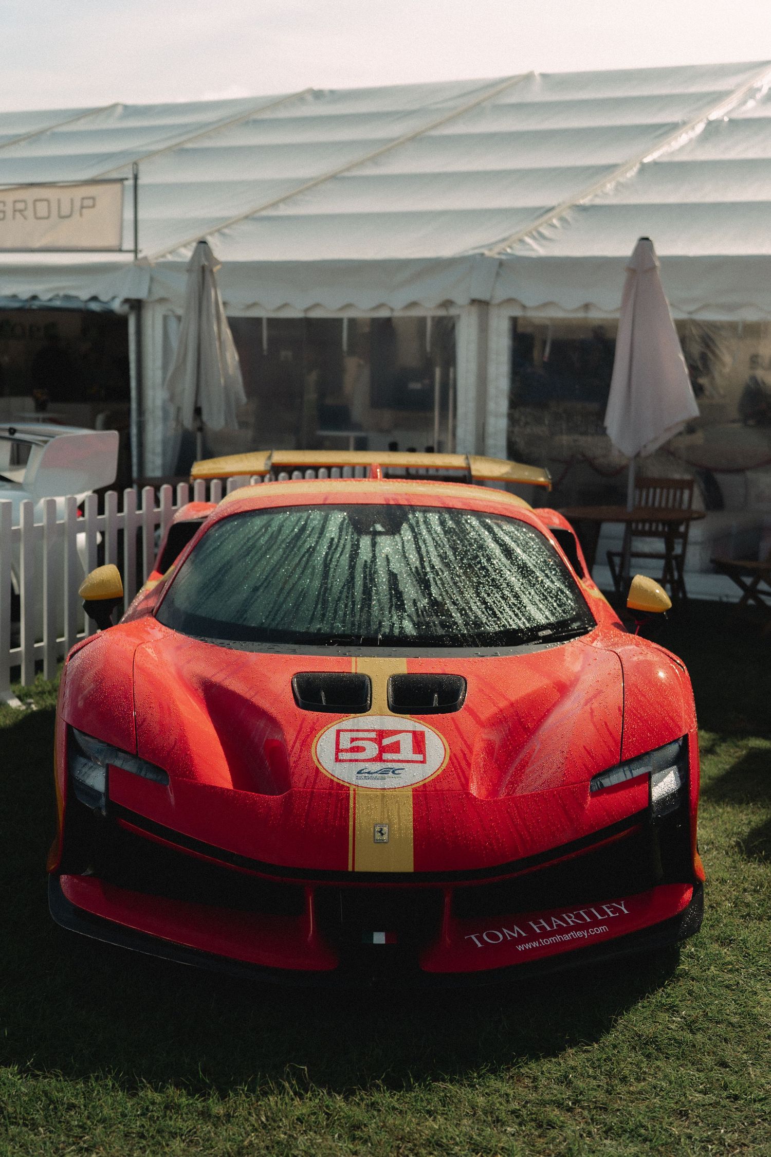 Red Ferrari 296 GT3 race car with number 51 livery displayed at Tom Hartley automotive event on grass.