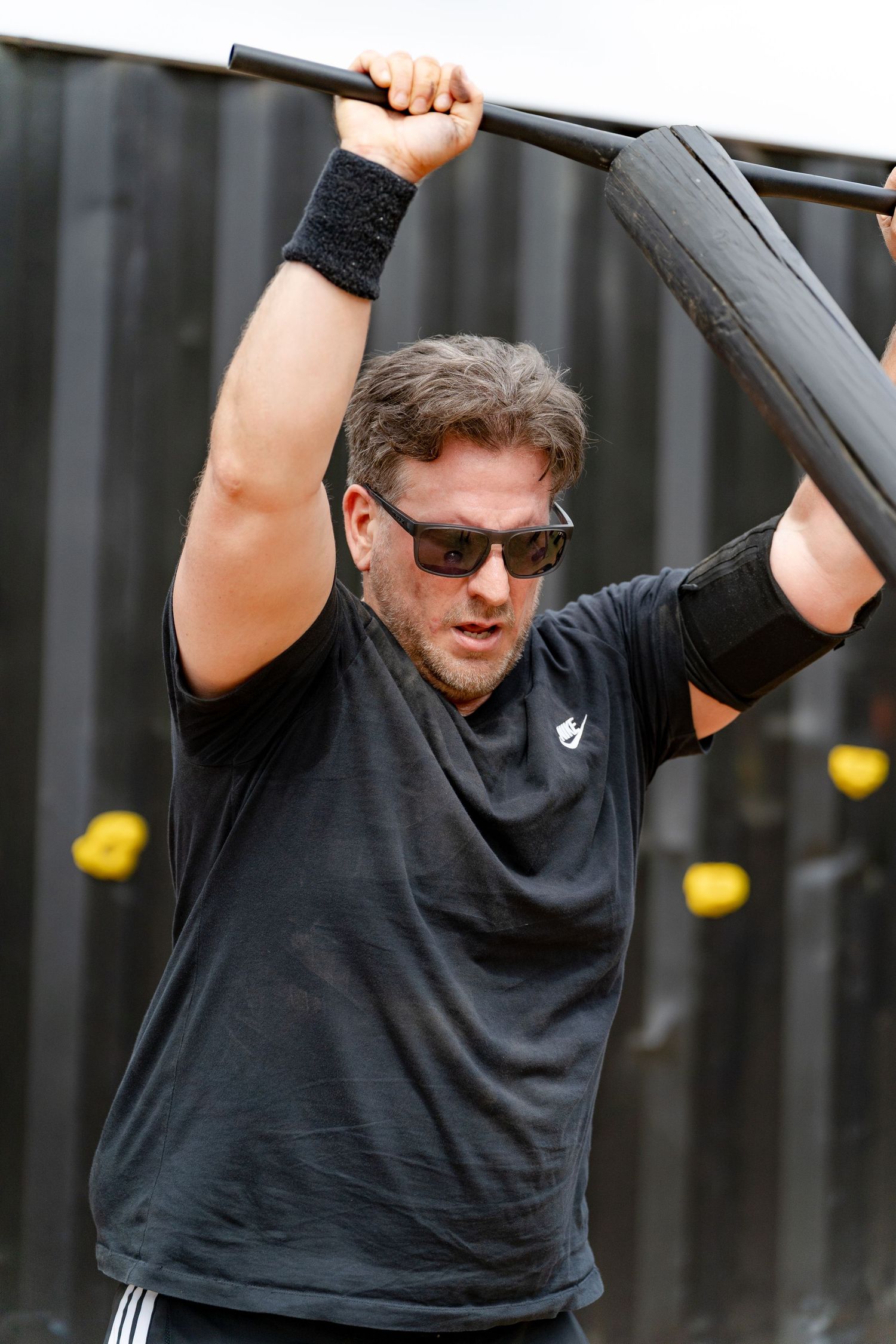 A muscular man in black shirt and sunglasses grips an overhead bar during an outdoor fitness challenge.