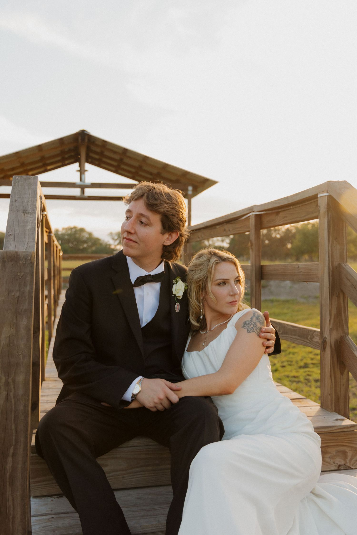 Bride and groom sitting on wooden deck at sunset, groom in black tuxedo, bride in white gown leaning on his shoulder.