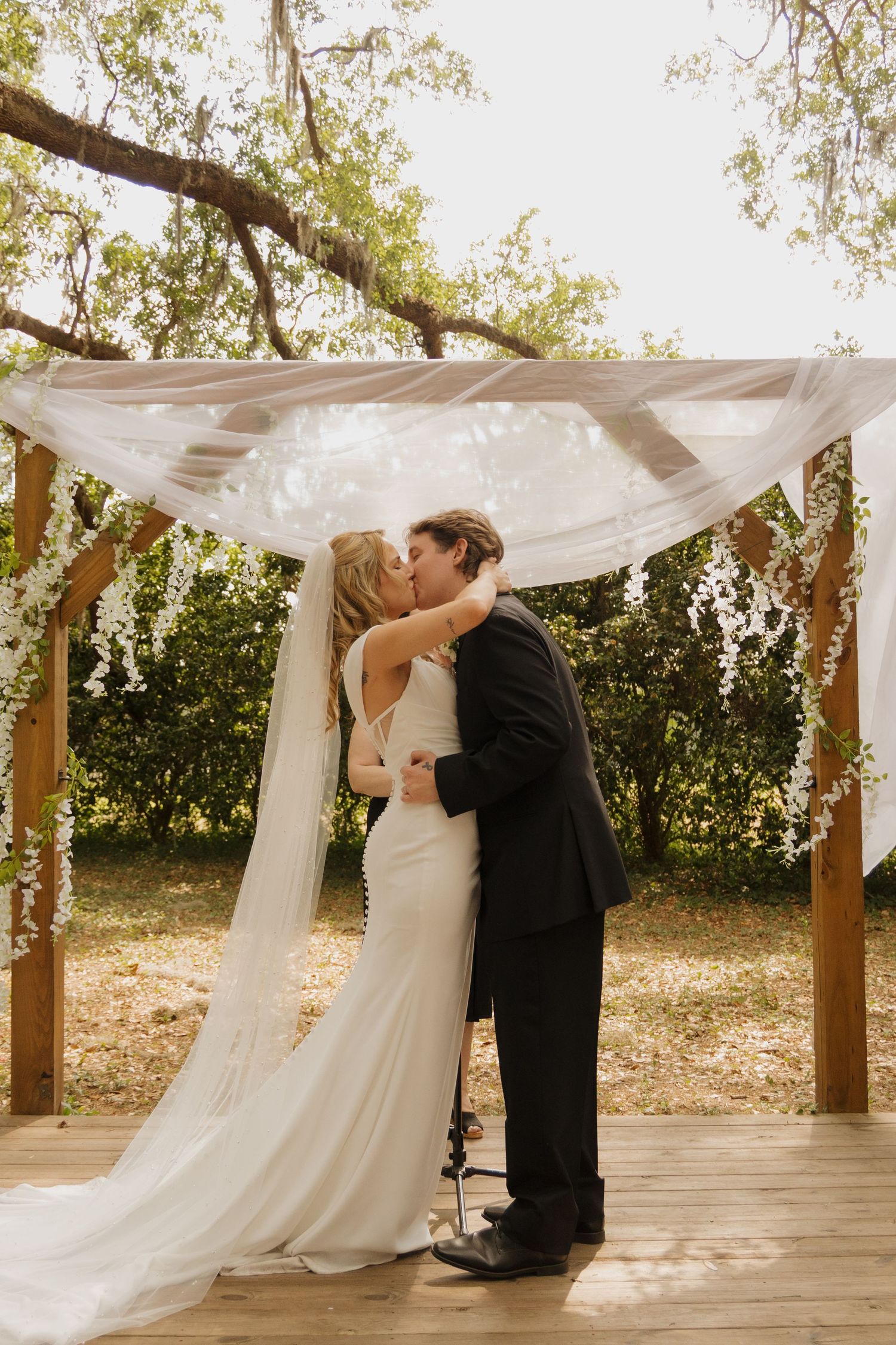 Bride and groom share first kiss under floral wooden arch with white draping at outdoor wedding ceremony.