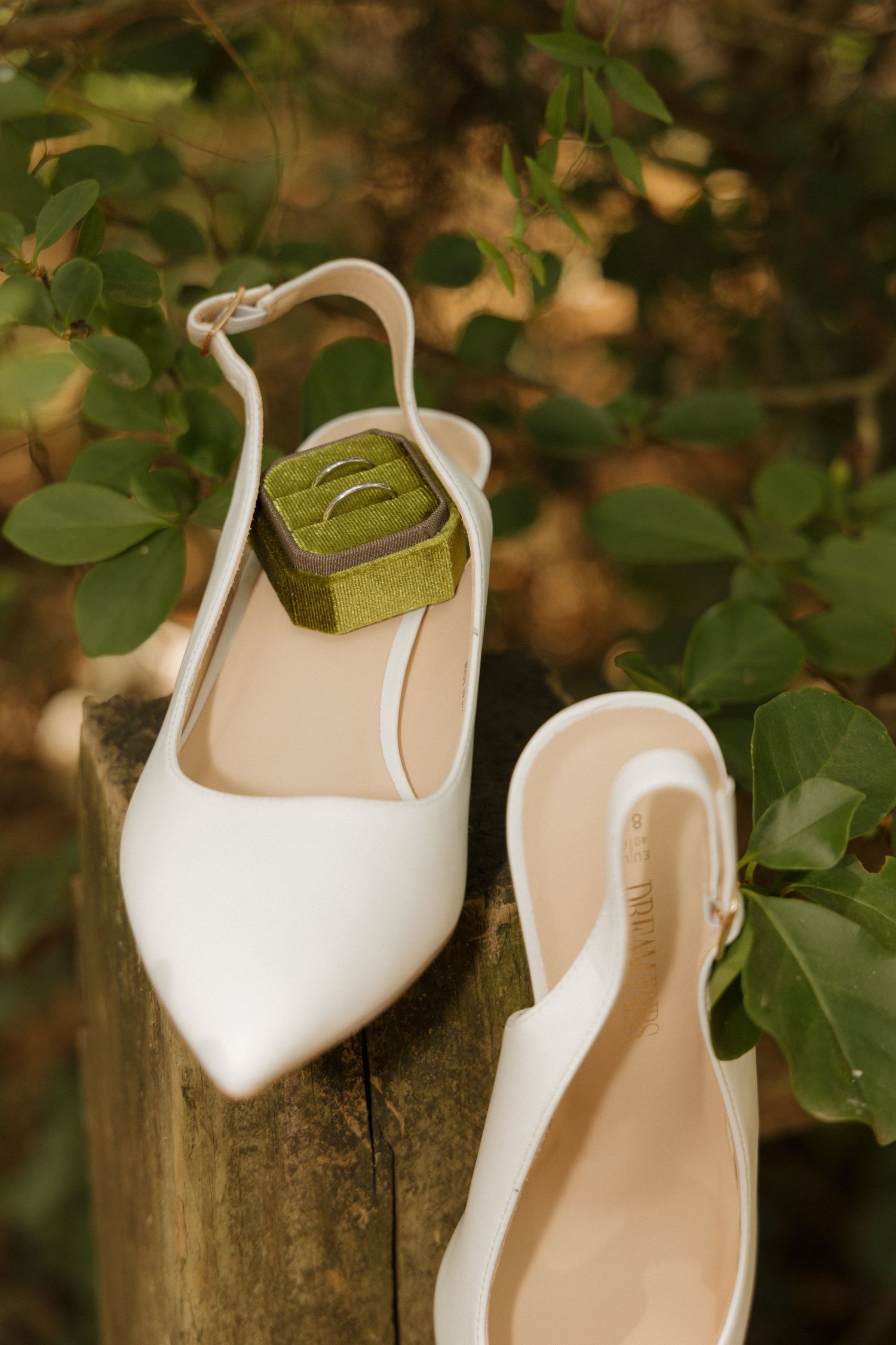 White slingback bridal heels with a green velvet ring box holding wedding bands, displayed on a wooden post outdoors.