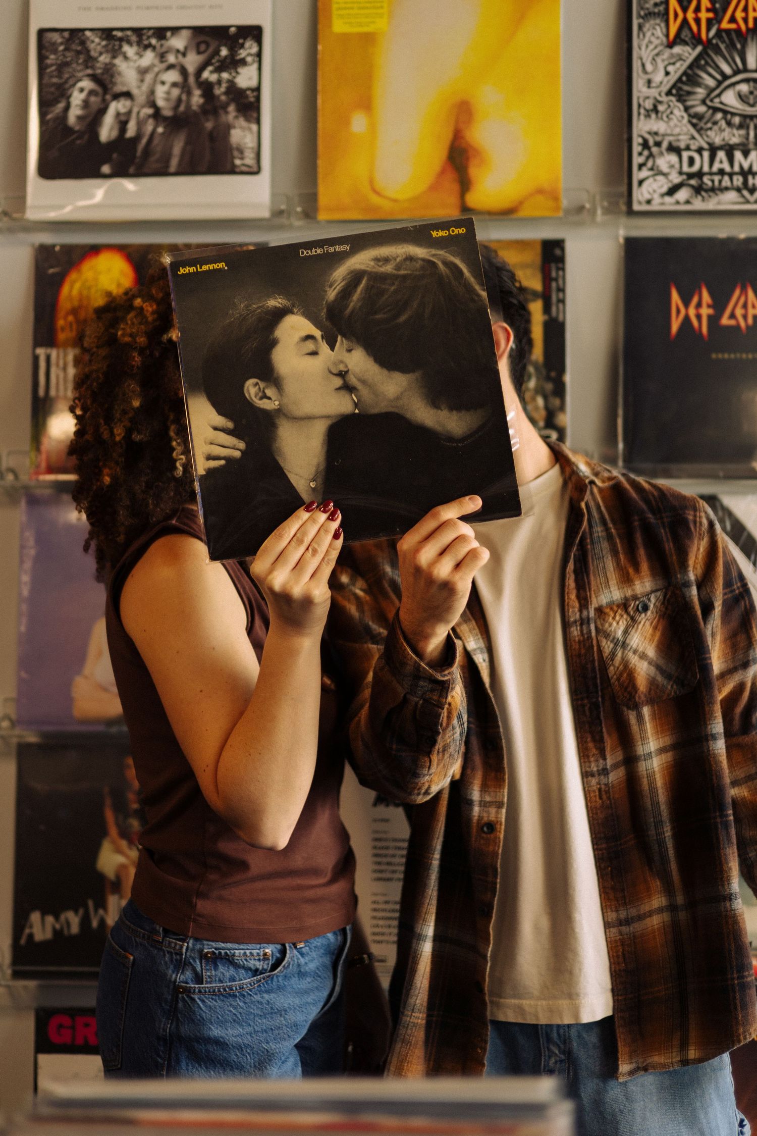 A couple embraces and kisses in front of a wall covered with music posters and album artwork.