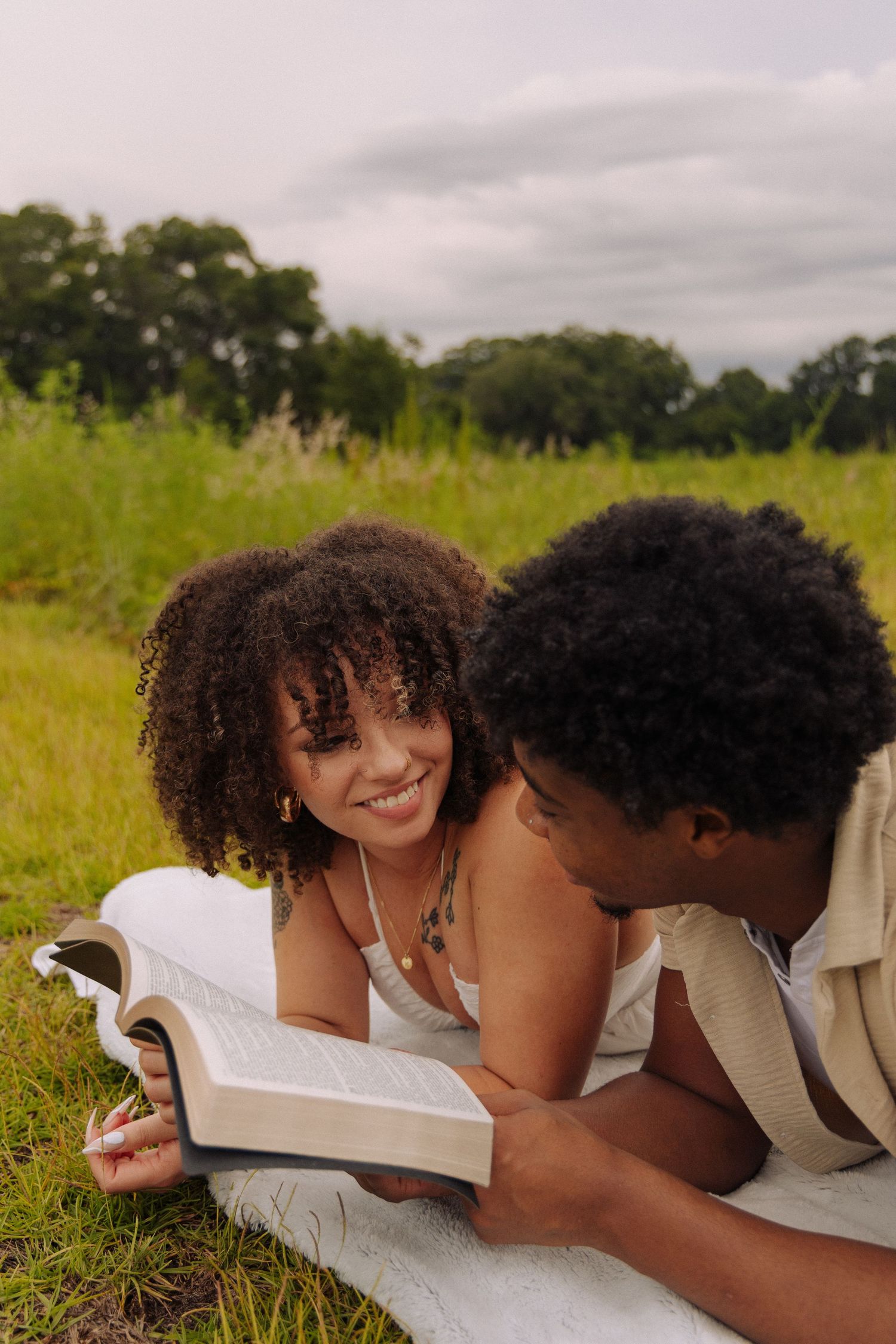 Two people share a tender moment while reading together on a blanket in a grassy field.