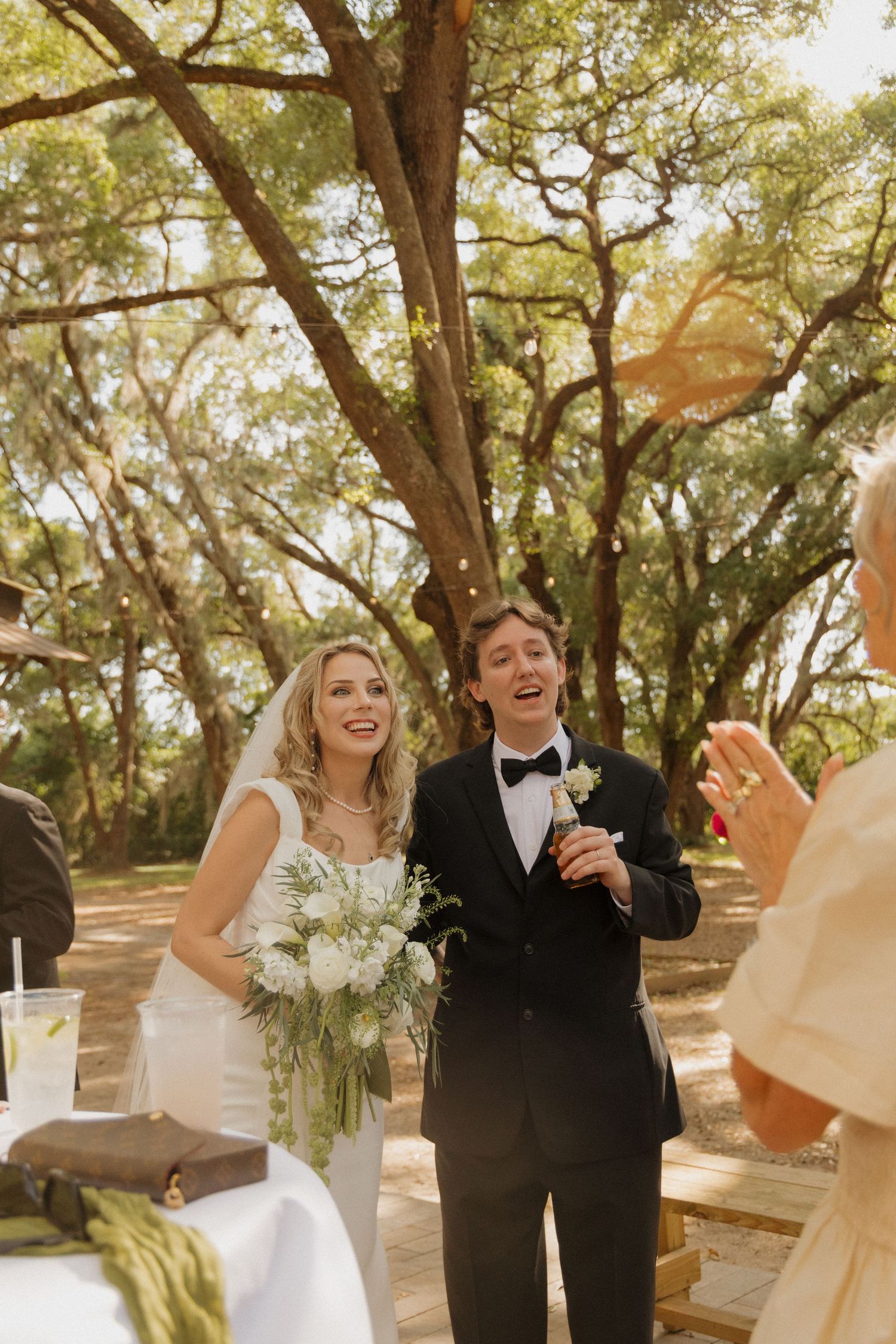 Joyful bride and groom laughing outdoors at wedding ceremony under oak trees, bride holding white floral bouquet.