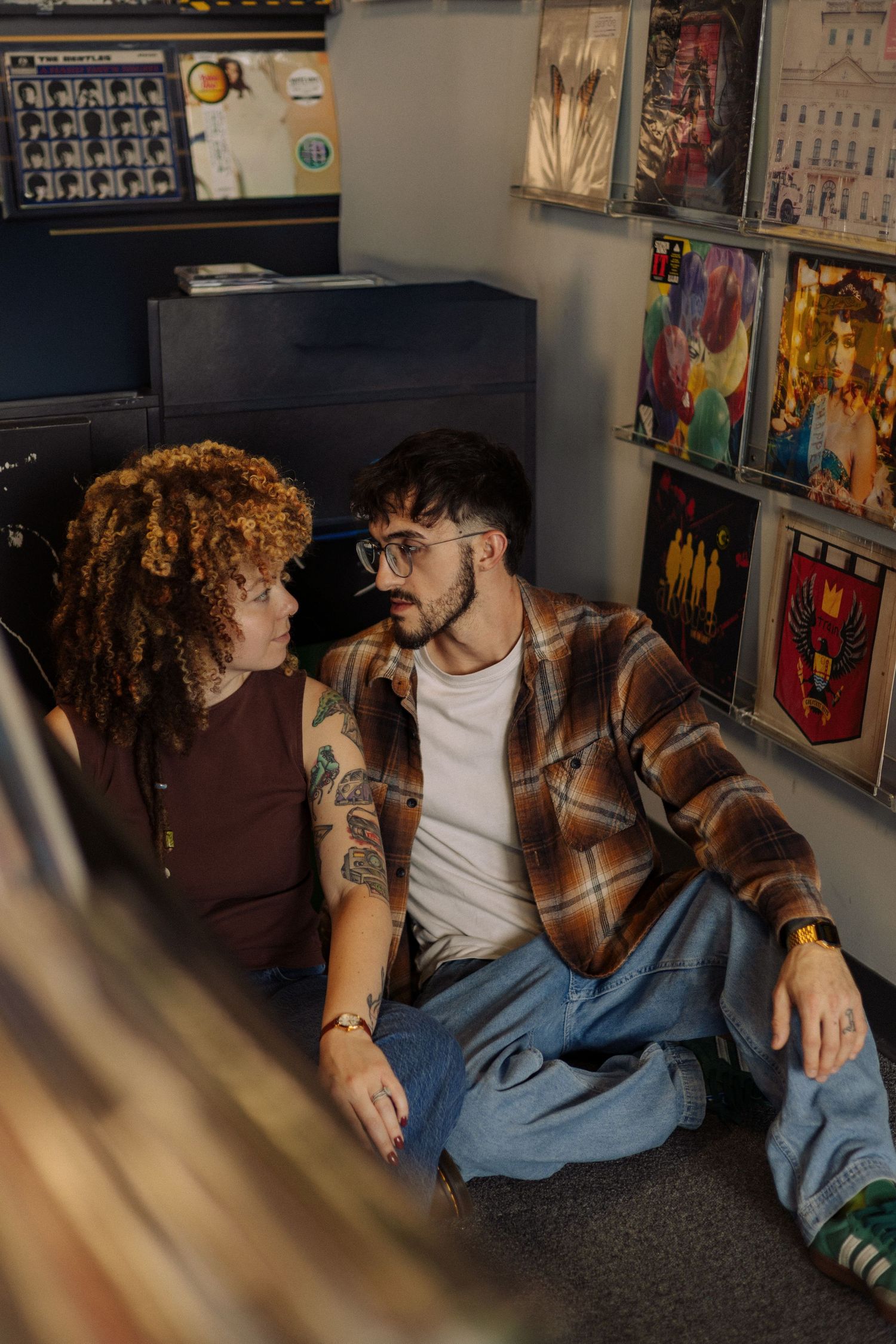 Two people in casual attire sit close together on the floor against a wall decorated with artwork and posters.