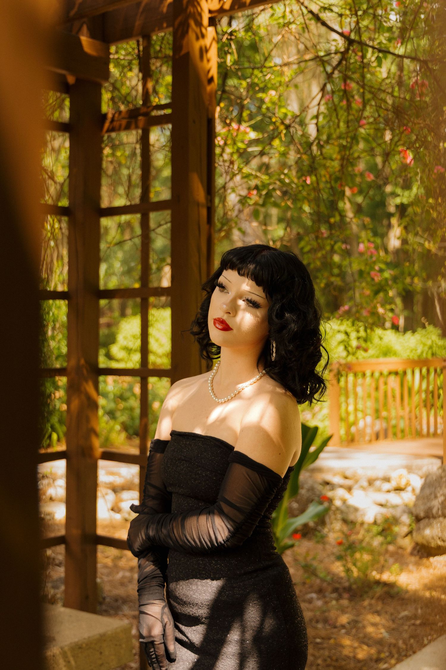 Woman in black off-shoulder gown and gloves poses gracefully in a sunlit garden with wooden pergola and lush greenery.