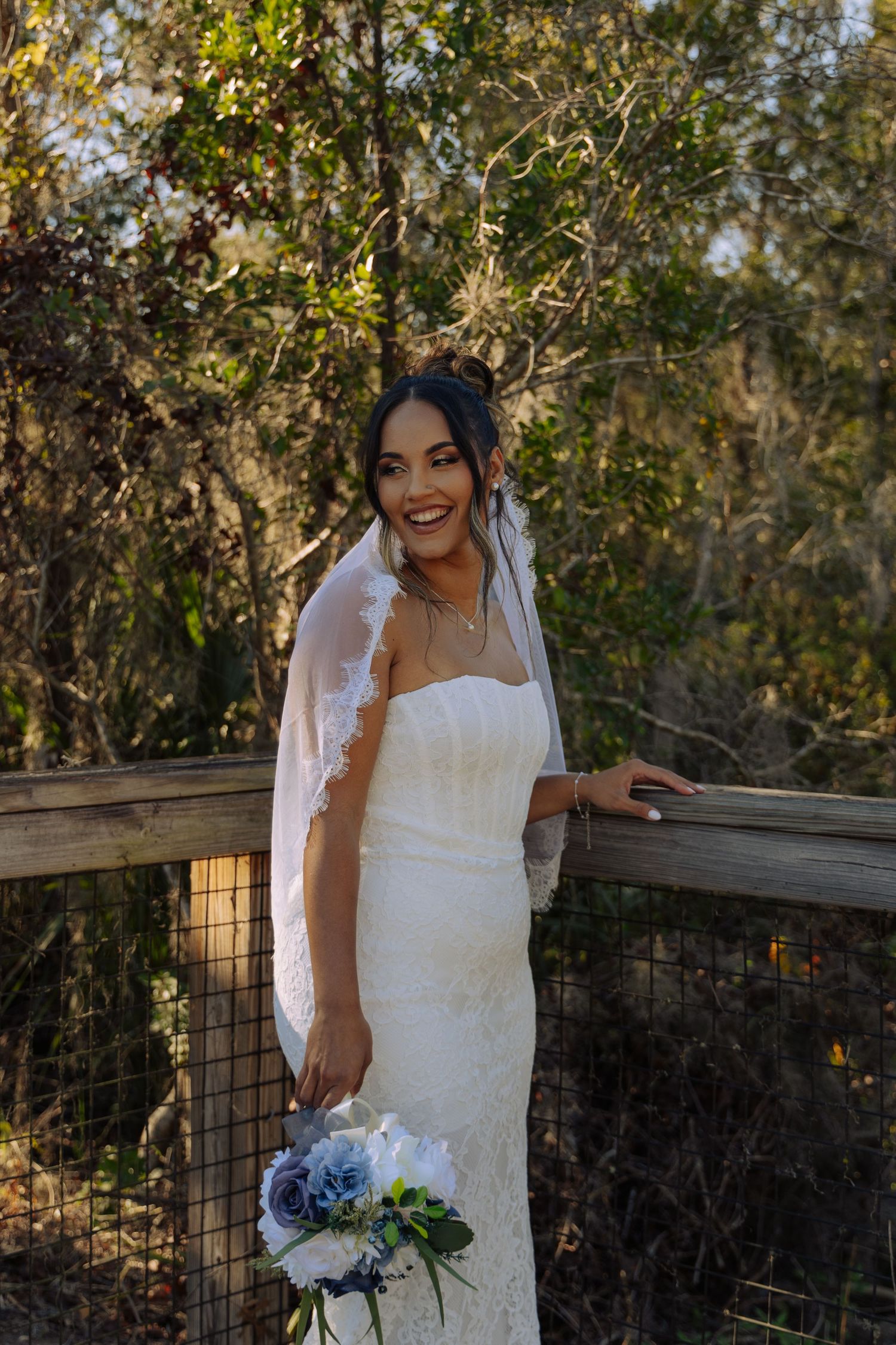 Bride in white dress poses joyfully against natural backdrop with floral bouquet.