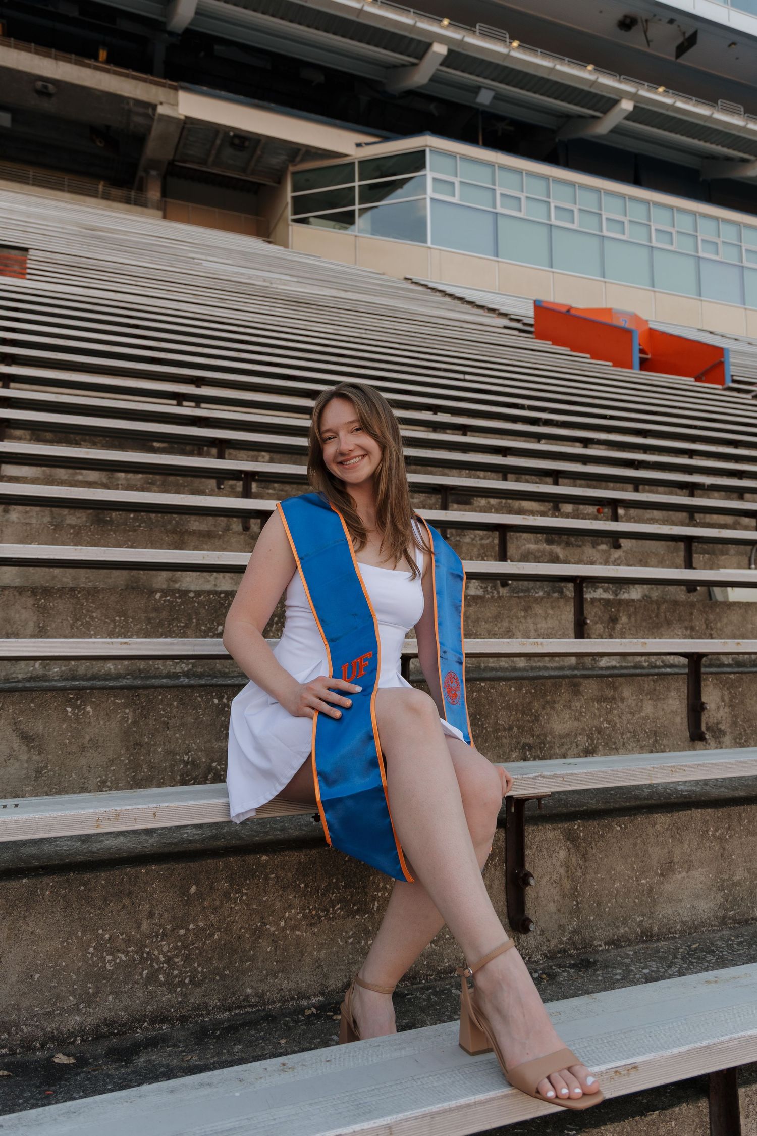 A smiling graduate in a white dress and blue sash sits on stadium bleachers for a celebratory senior portrait.