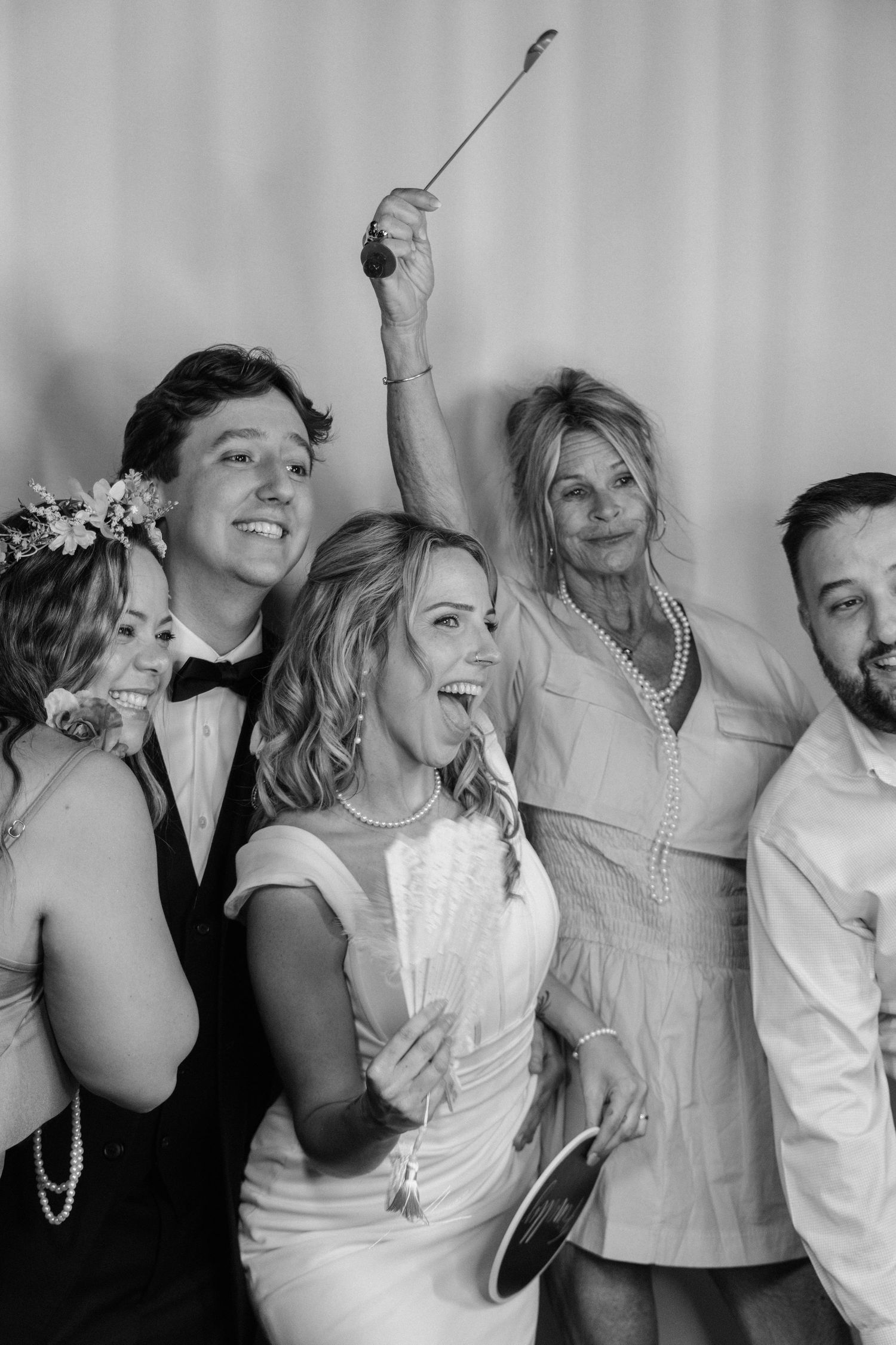 Black and white wedding photo booth shot of bride in white dress with groom in tuxedo and joyful guests holding props.