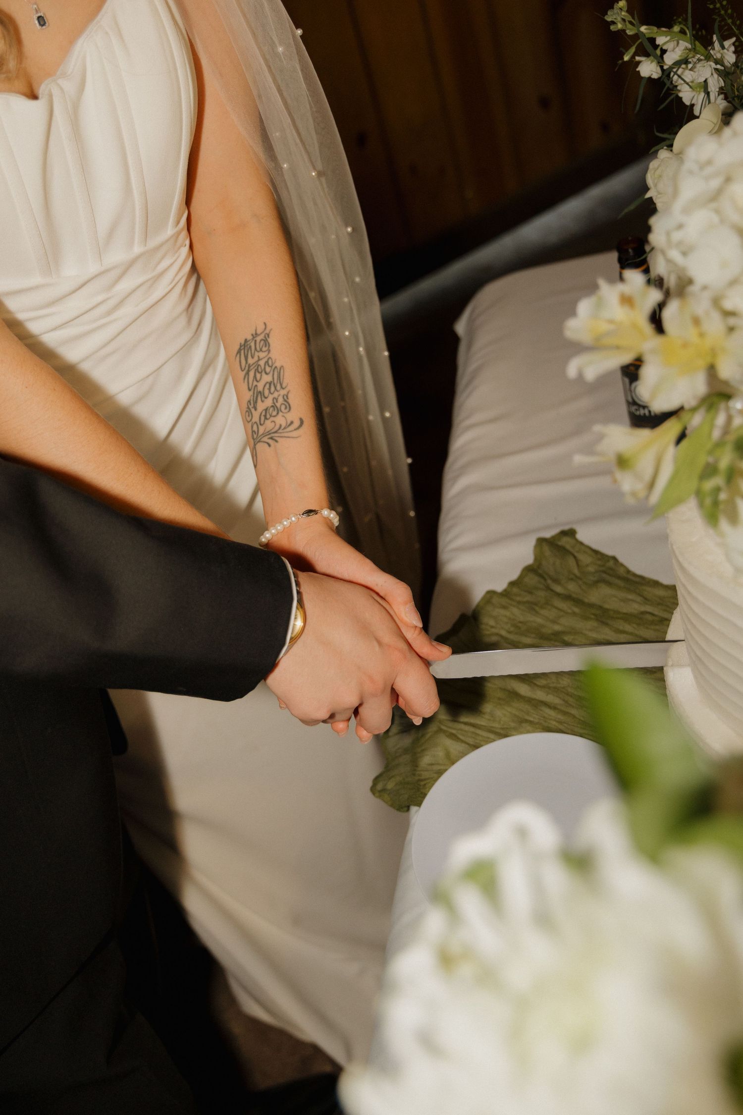Bride and groom cutting white wedding cake together, bride wearing pearl bracelet with forearm tattoo visible.
