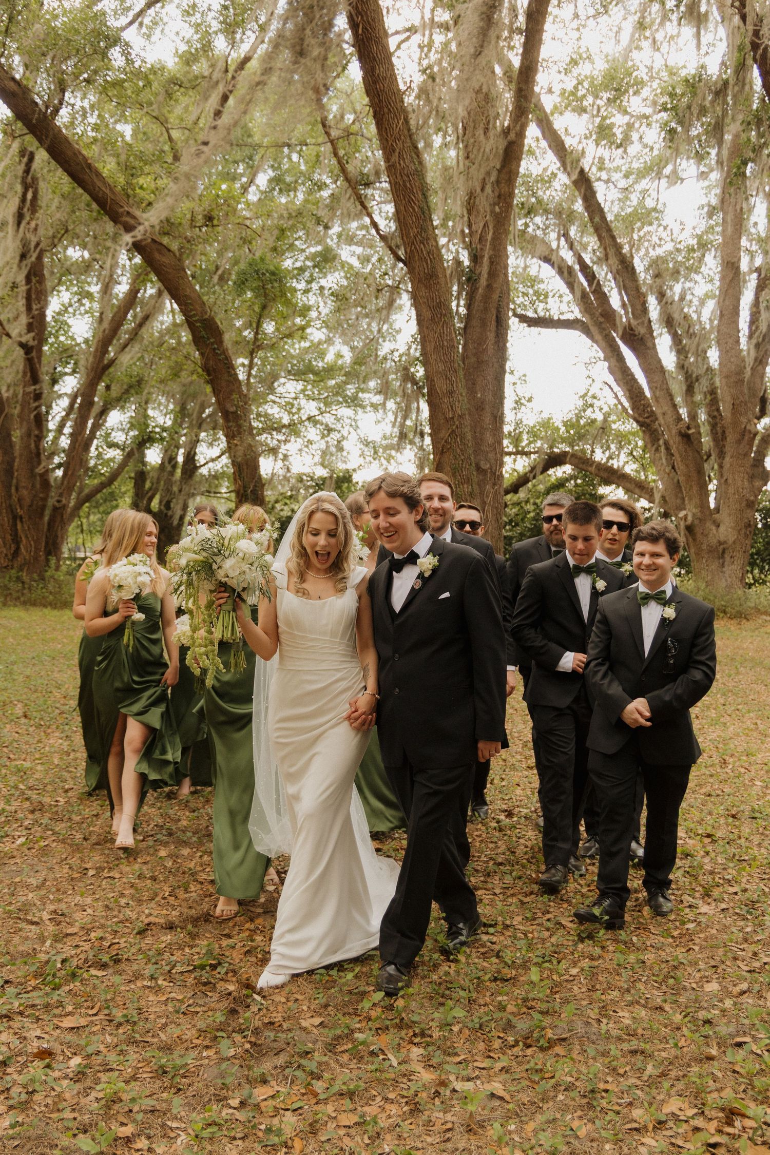 Wedding party walks outdoors under moss-draped trees, bride in white gown with bouquet, groom in tuxedo, bridesmaids in sage green.
