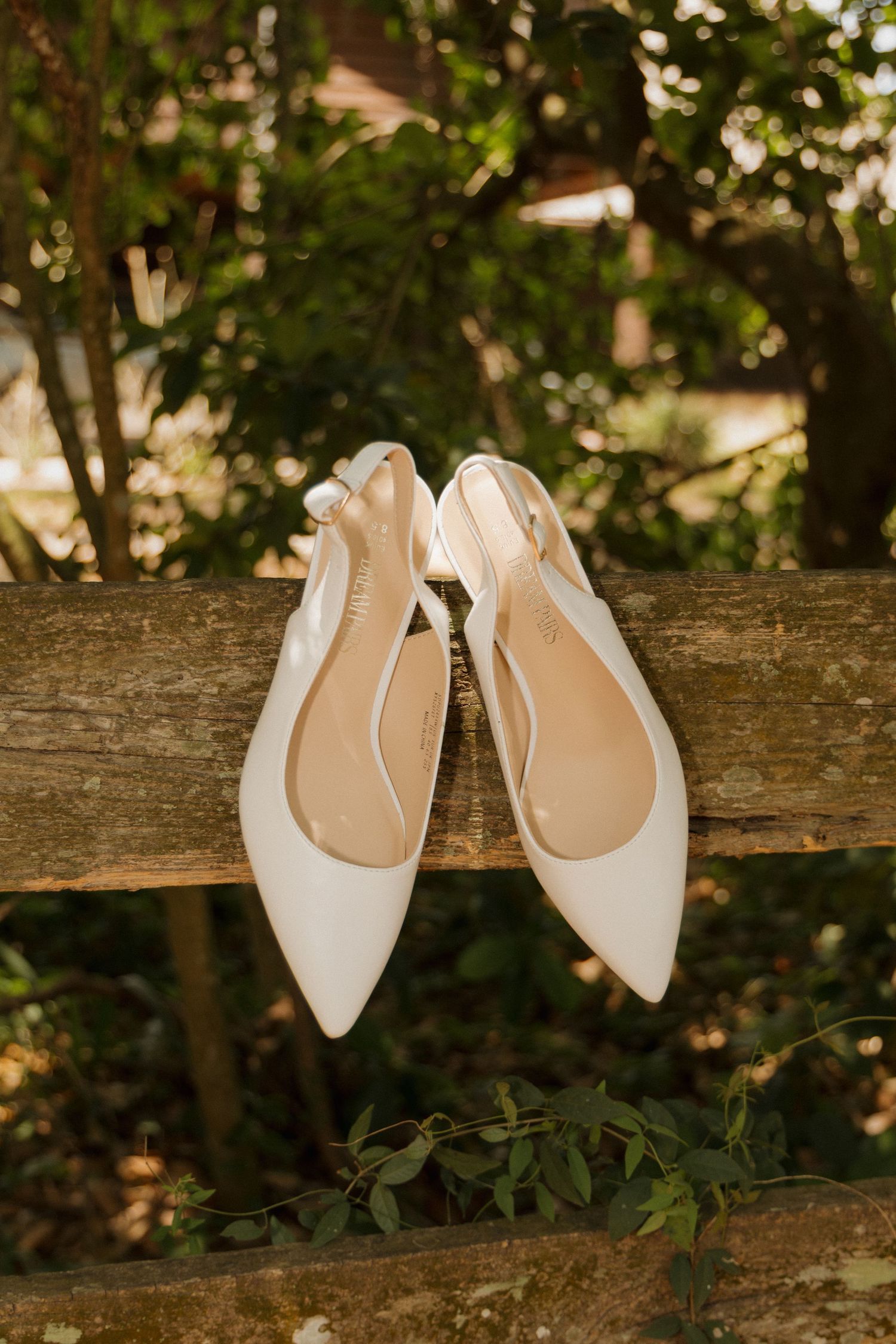 White slingback pointed-toe bridal flats resting on a rustic wooden fence surrounded by lush green foliage.
