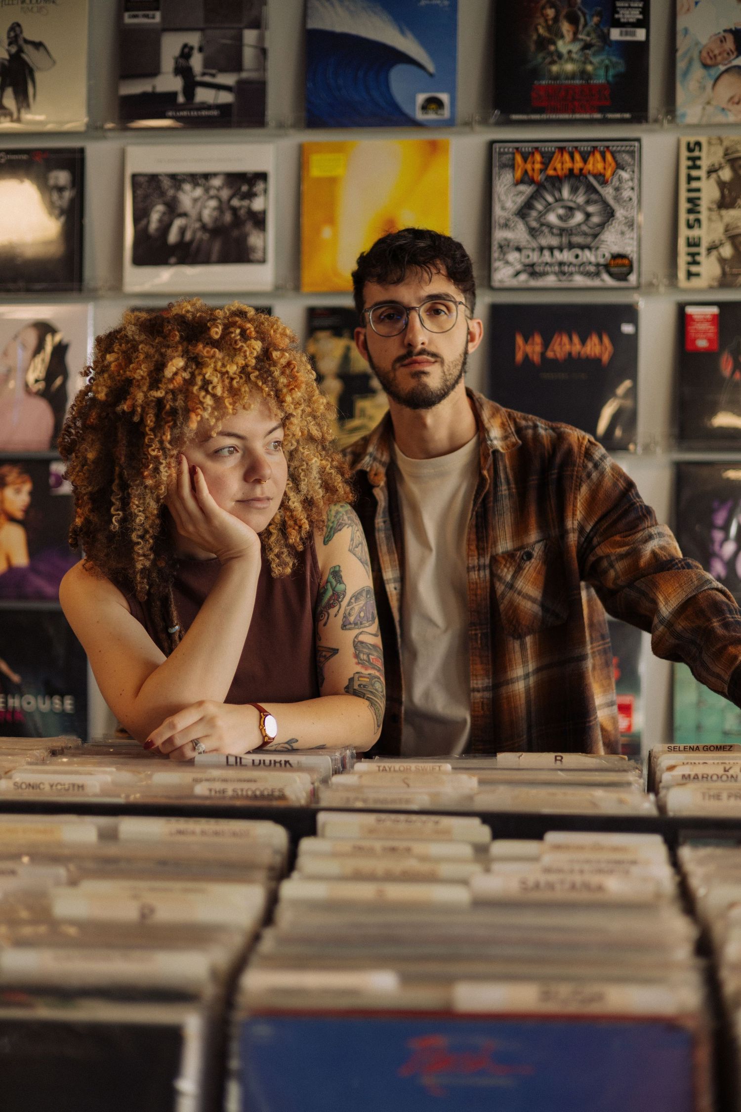 Two people browsing vinyl records in a music store with album covers and posters displayed on the wall behind them.