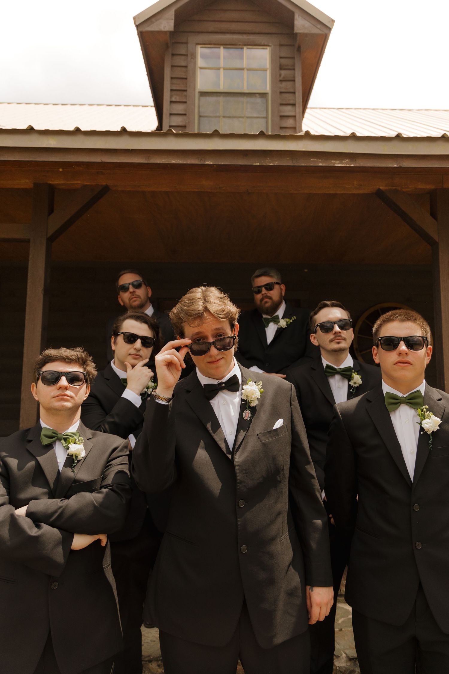 Groomsmen in black tuxedos and green bow ties wearing sunglasses pose coolly outside a rustic wooden building.