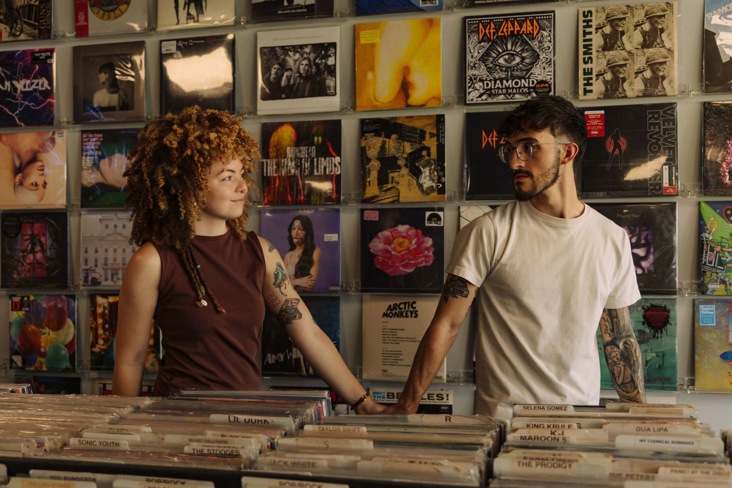 Two people browse through vinyl records in a music store with album covers displayed on the wall.