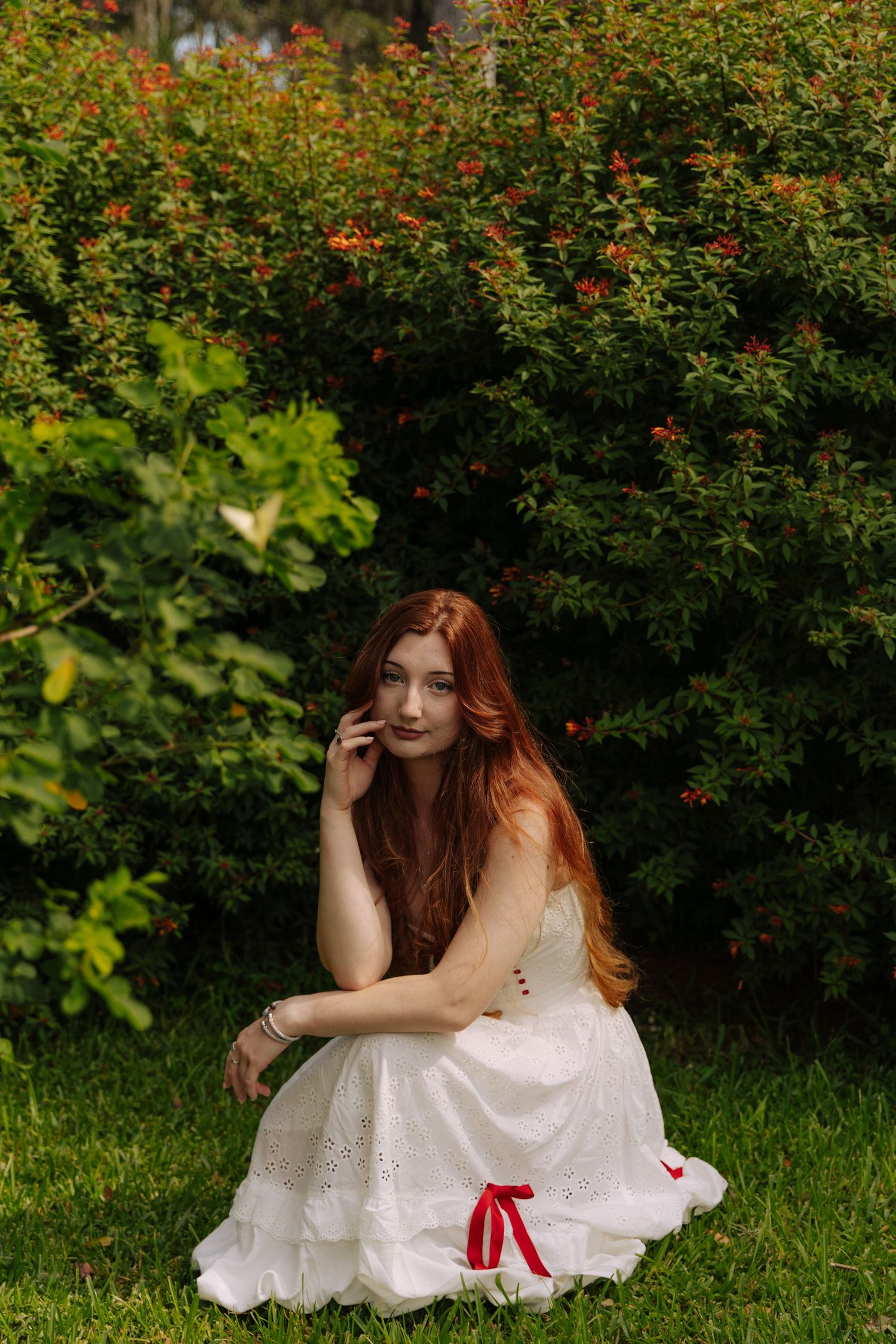 Person in white dress poses gracefully near flowering bushes in garden setting.