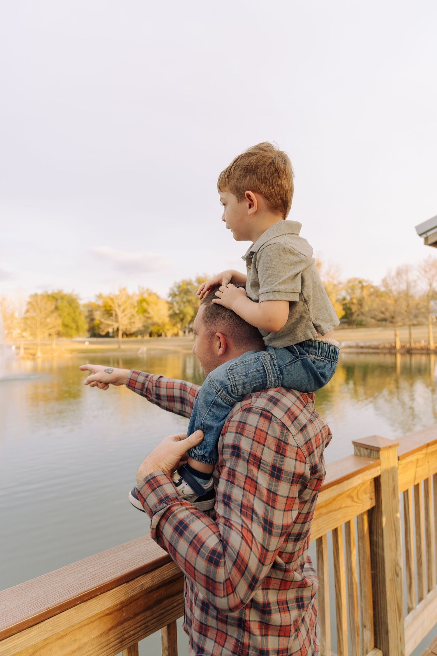 A child and adult in plaid shirt sit together on a wooden deck overlooking a peaceful lake at sunset.