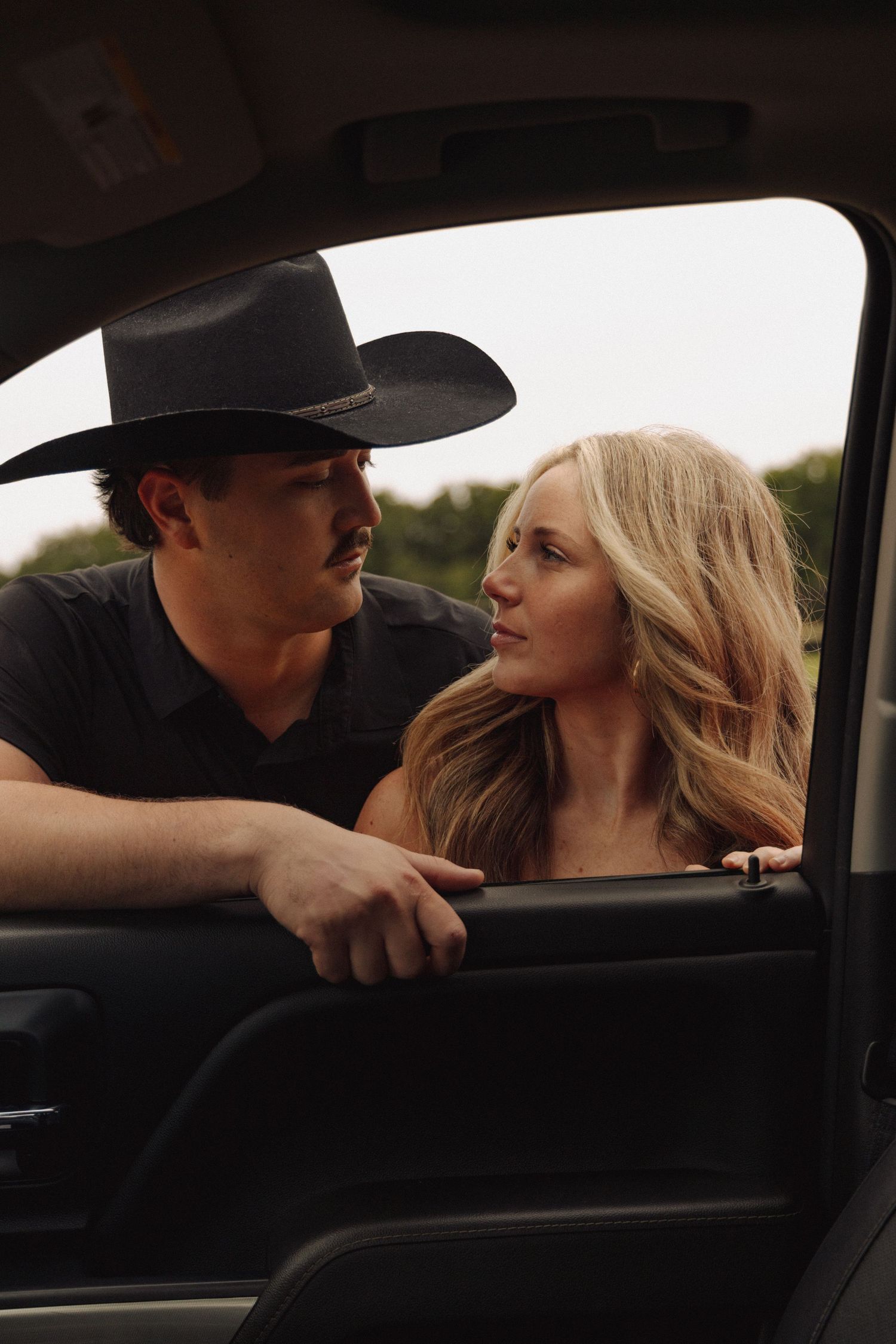 A couple shares an intimate moment inside a car with a cowboy hat visible in silhouette.