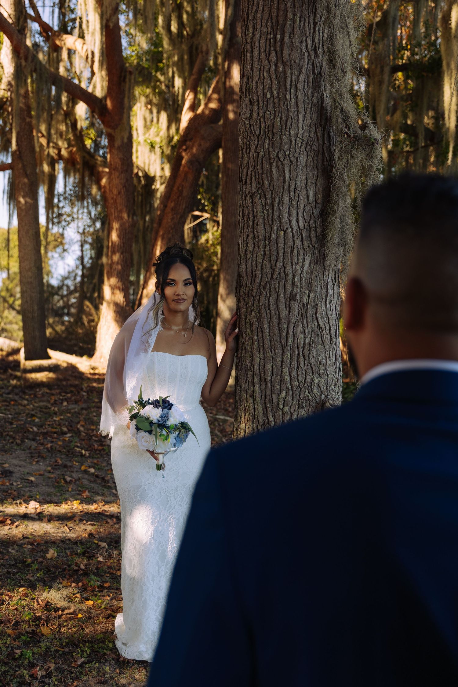 A bride in a white dress stands among tall trees in a wooded outdoor setting as sunlight filters through the branches.