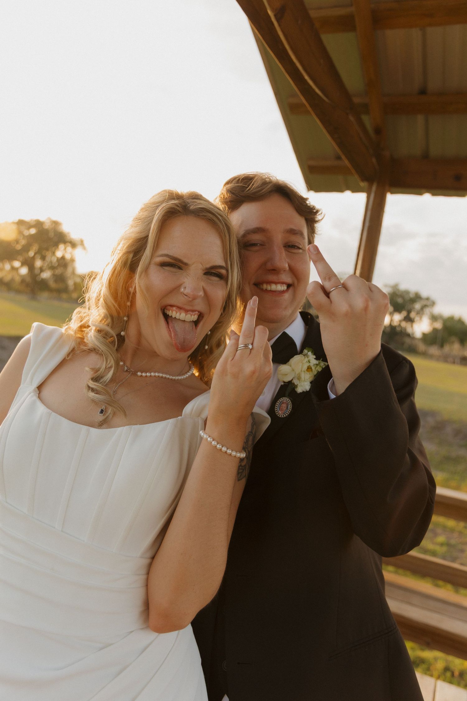 Joyful newlyweds showing off wedding rings, bride in white dress and groom in black suit, golden hour outdoor wedding photo.
