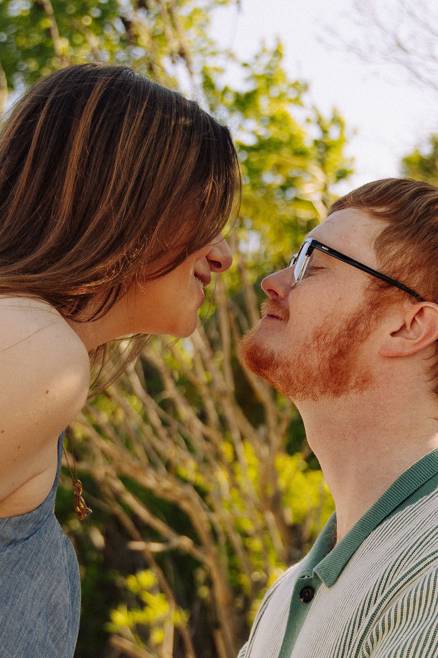 A couple sharing a romantic moment about to kiss outdoors, man with red beard and glasses, woman with brown hair, green trees behind.