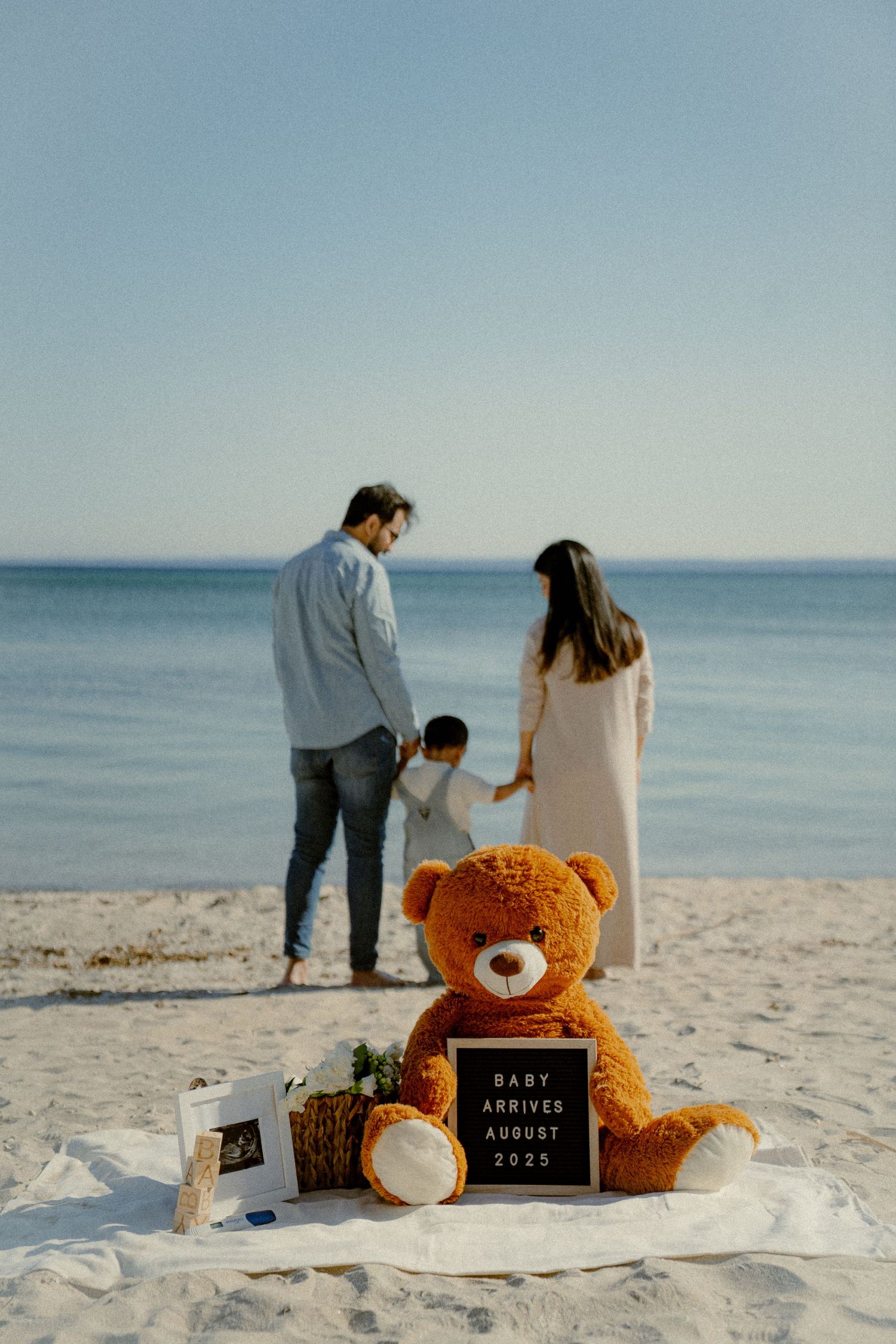 A teddy bear and letter board sit on beach sand while a family stands by the ocean.