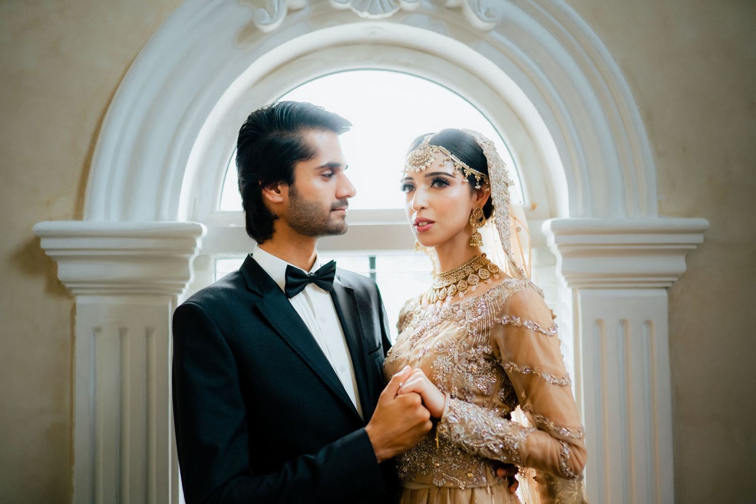 An elegant wedding couple poses under ornate white archways in traditional South Asian formal wear.