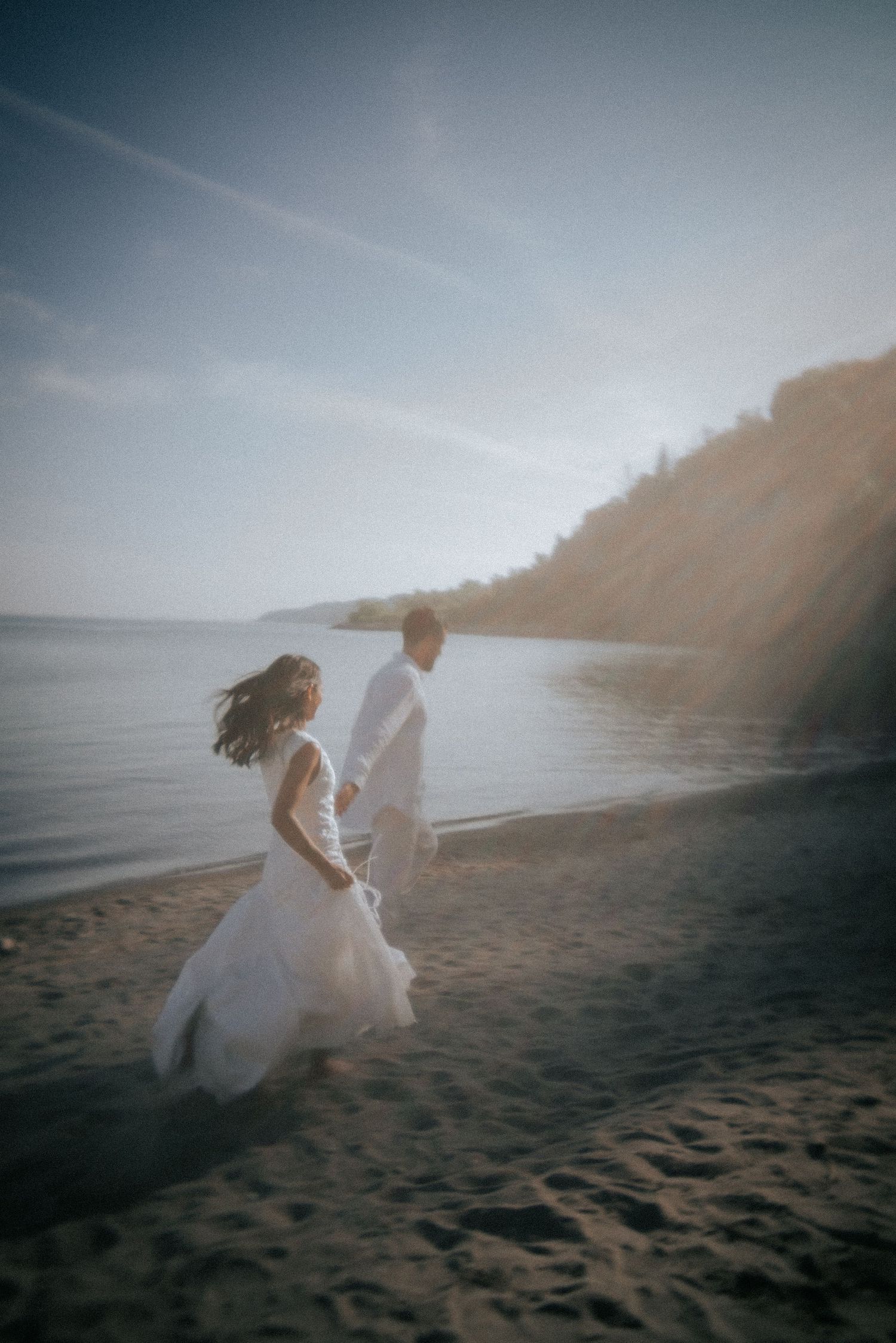 A couple in white outfits runs playfully along a misty beach at dusk.