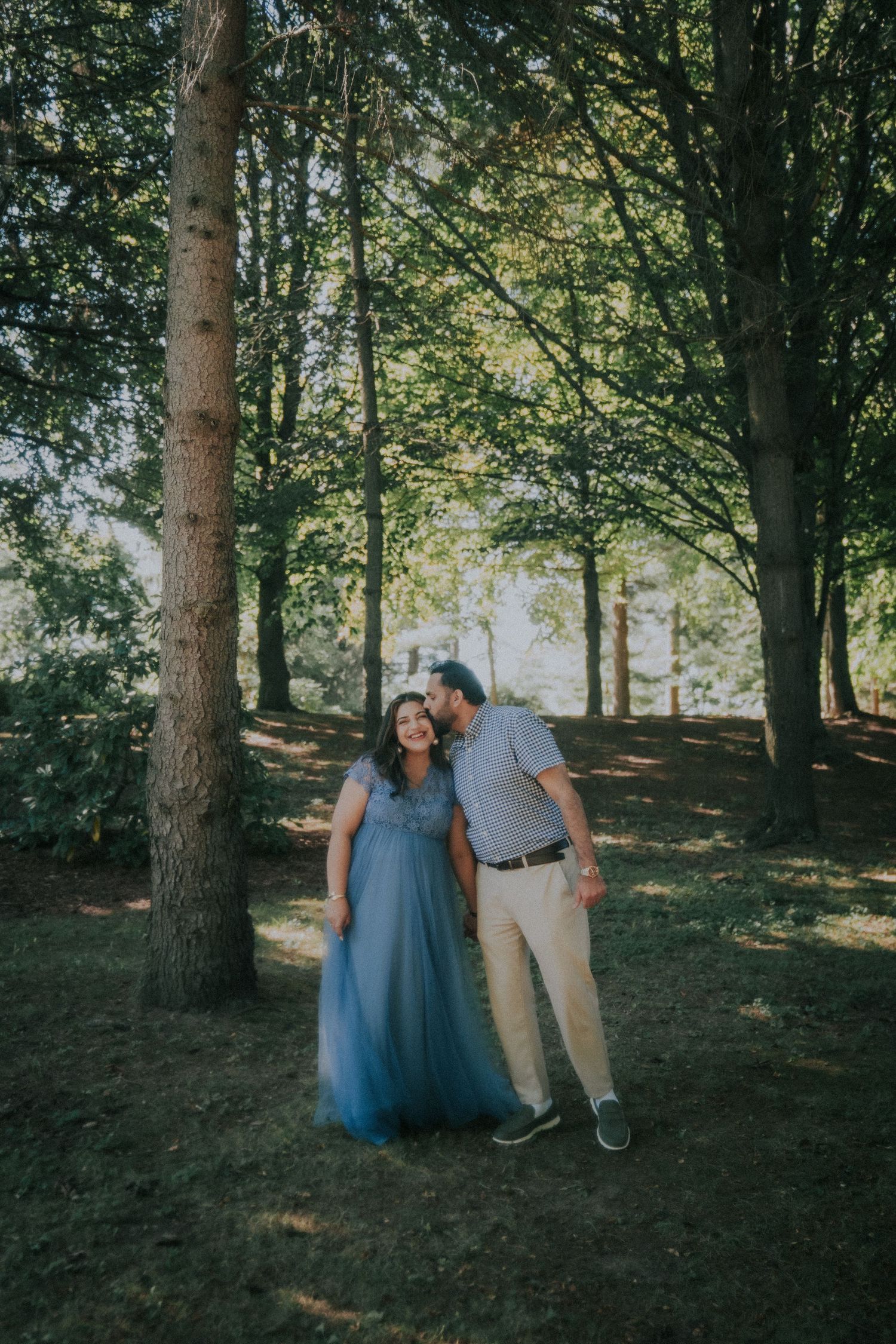A couple poses playfully among tall trees in a wooded forest setting.