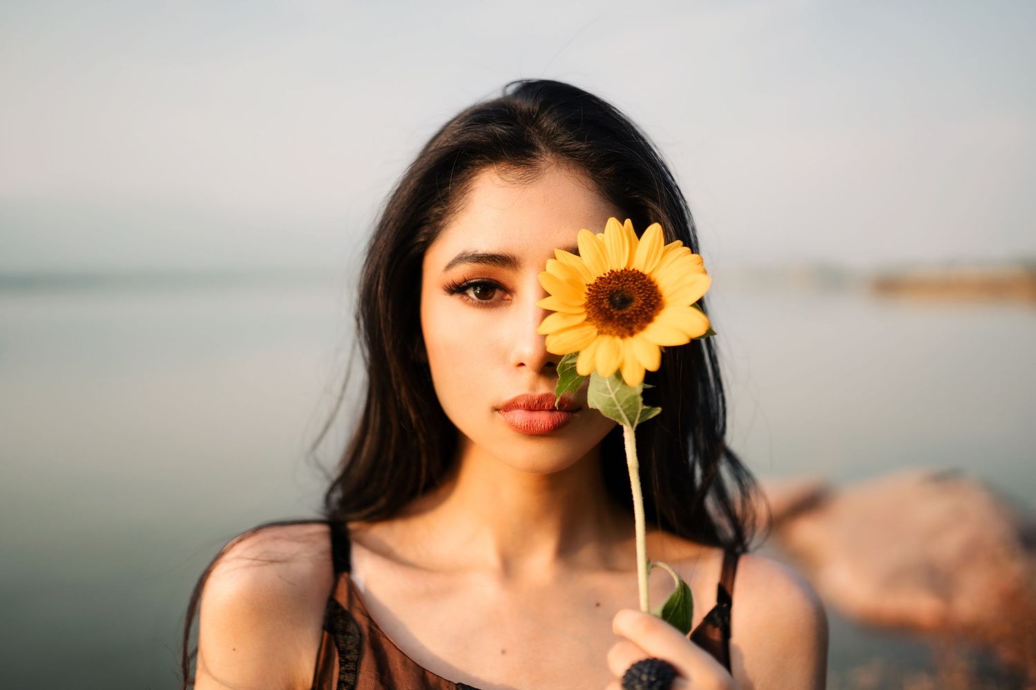 A person holds a bright yellow sunflower against a serene waterfront background.