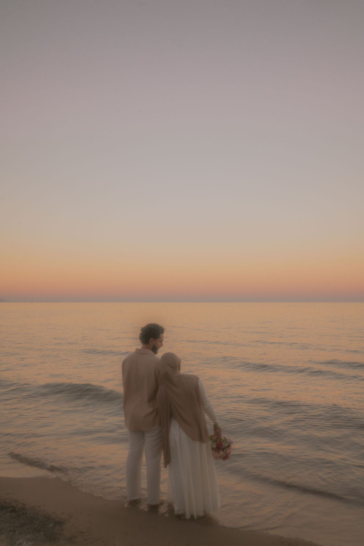 A couple walks along the beach during a dreamy sunset with soft pastel colors.