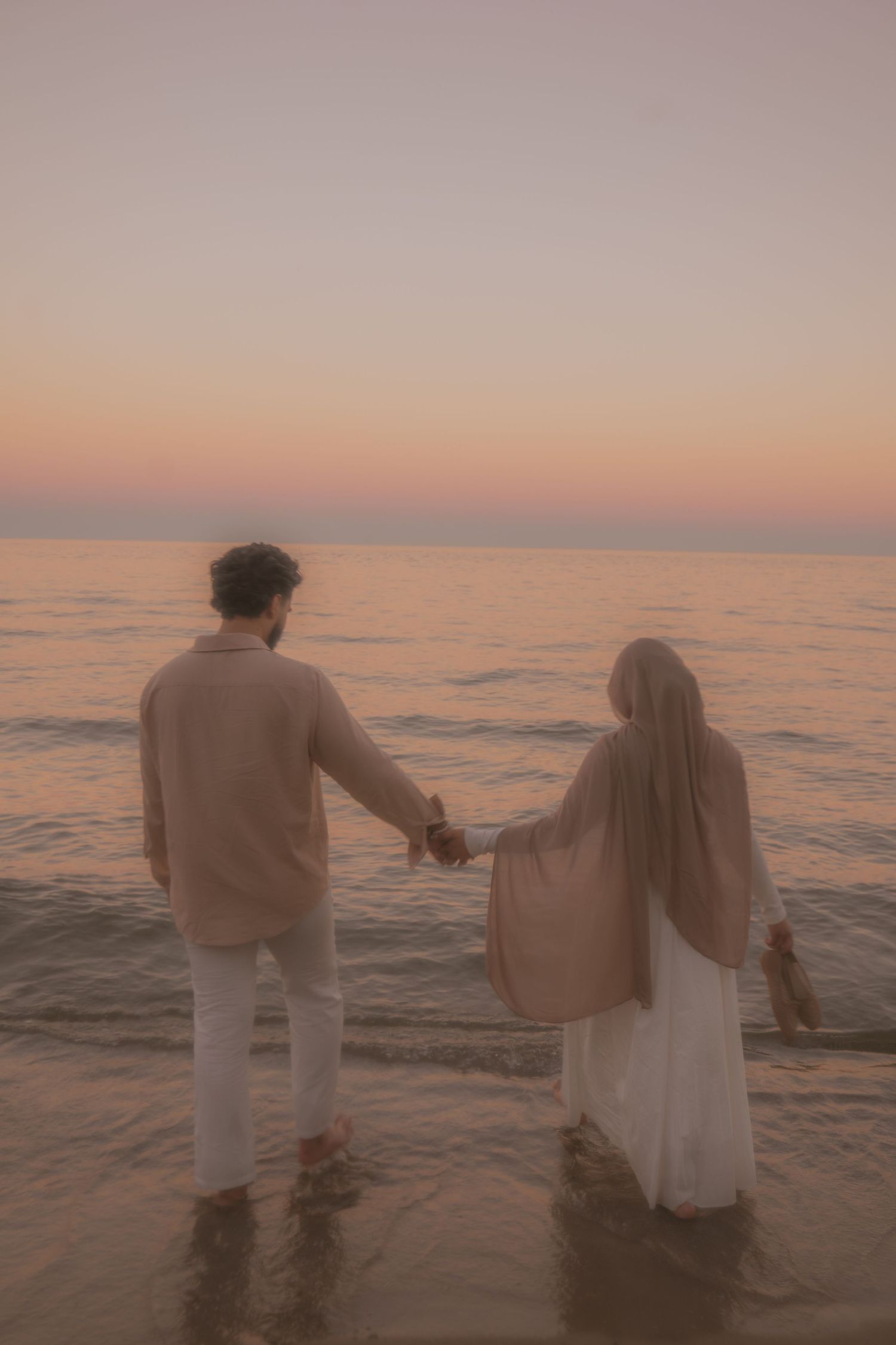 A couple holds hands while walking along the shoreline at sunset.