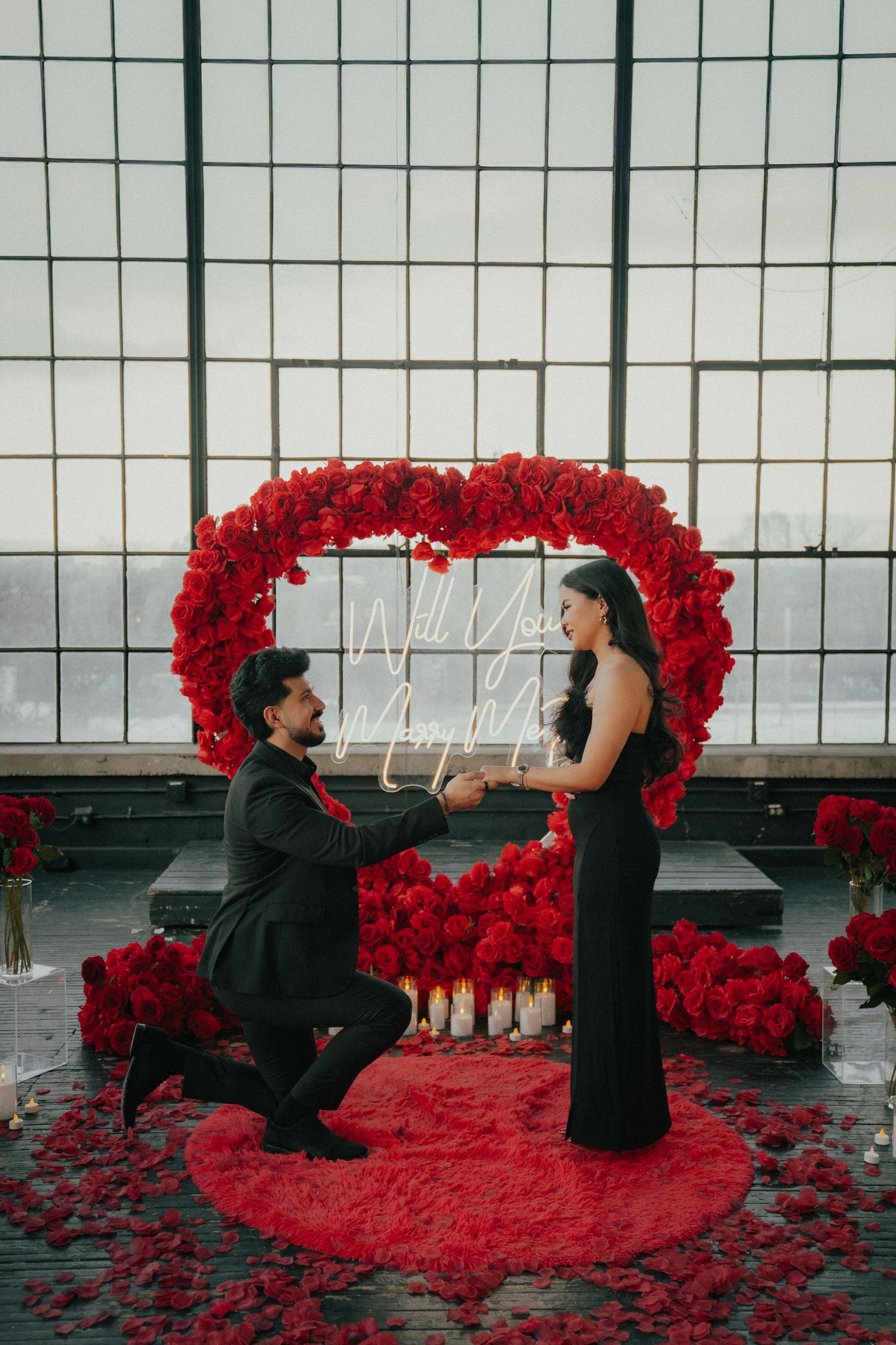 Romantic proposal scene with red rose petals arranged in a heart shape against a modern window backdrop.
