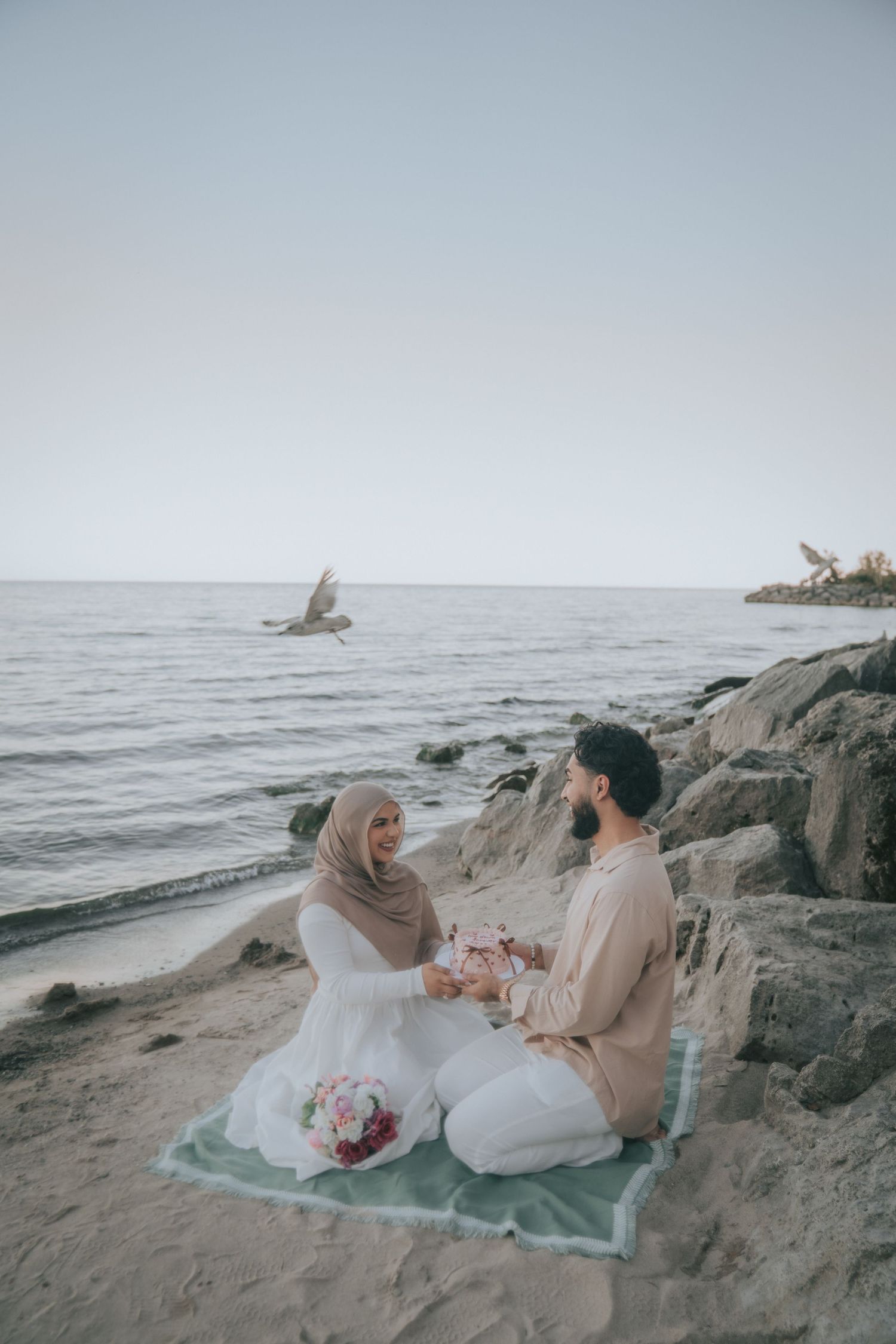 A couple relaxes on rocks by the beach with a picnic blanket and flowers.