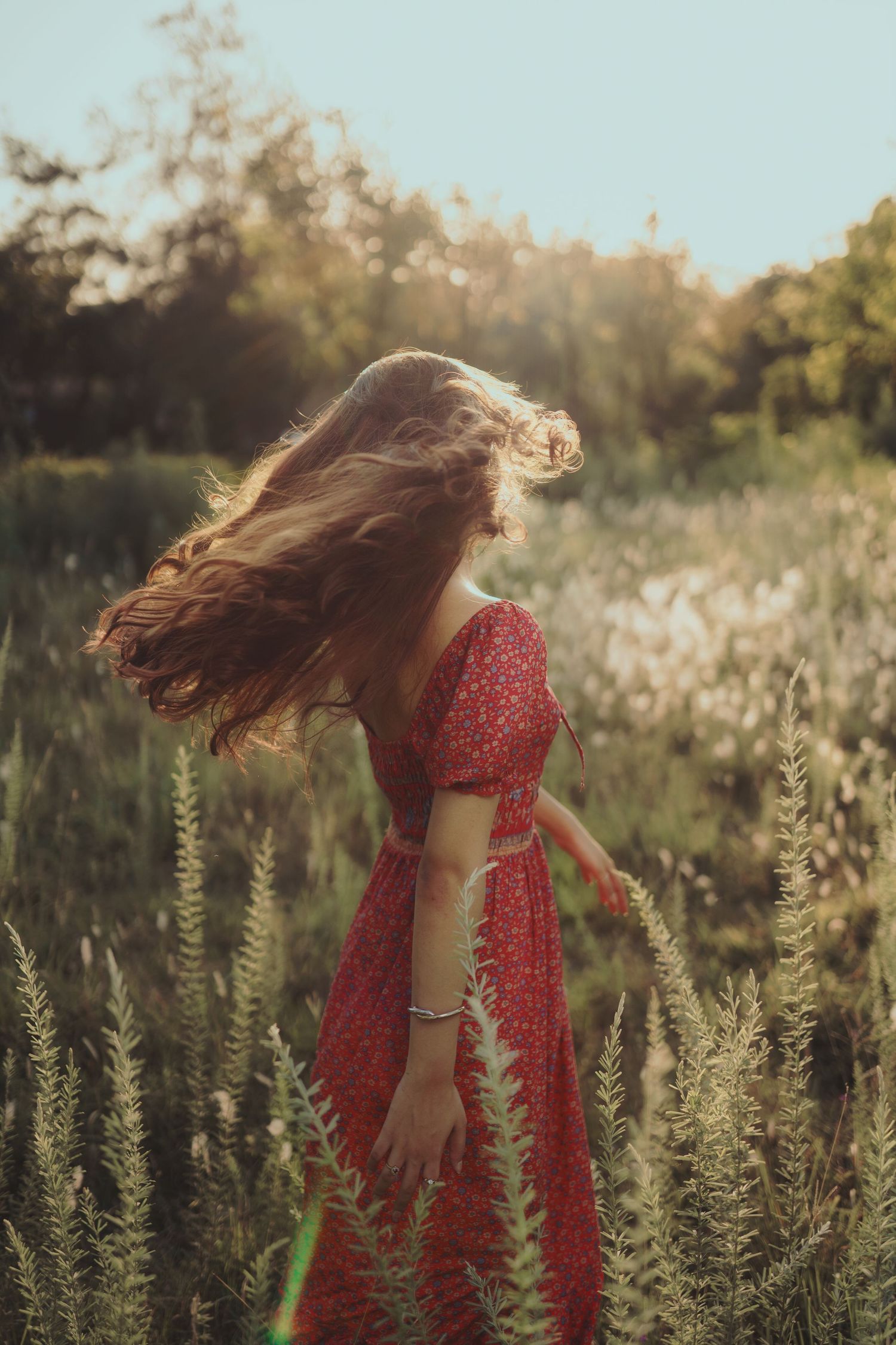 Long hair flows in sunlight as figure in red dress stands among wild summer grasses.