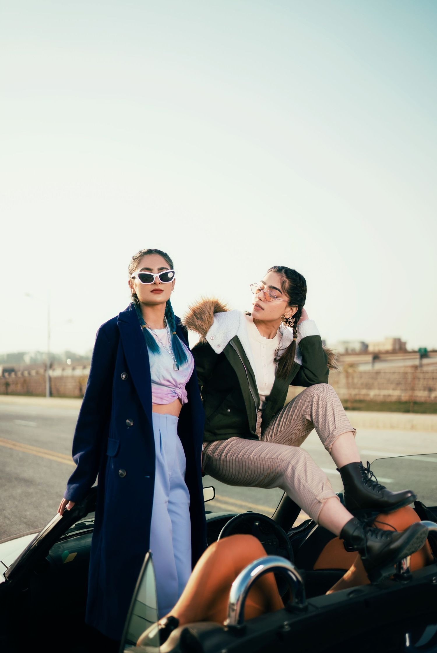 Stylish figure poses on motorcycle wearing navy coat and sunglasses in urban setting.