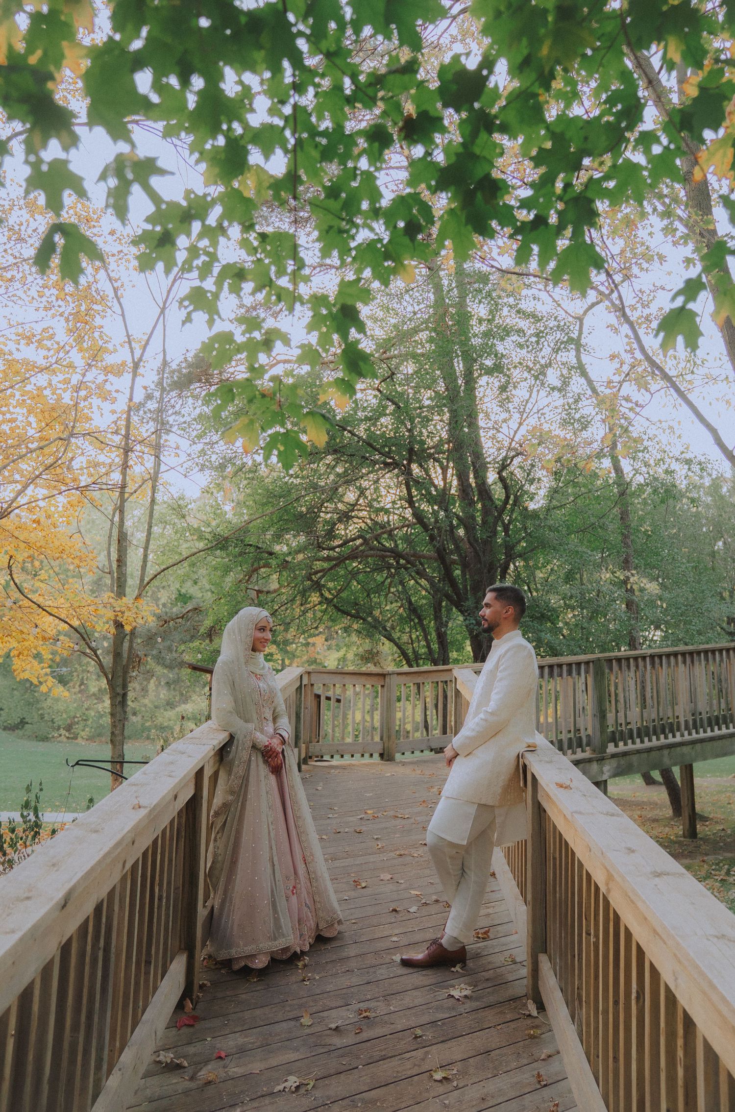 Romantic couple in vintage attire walking along wooden boardwalk surrounded by autumn foliage.