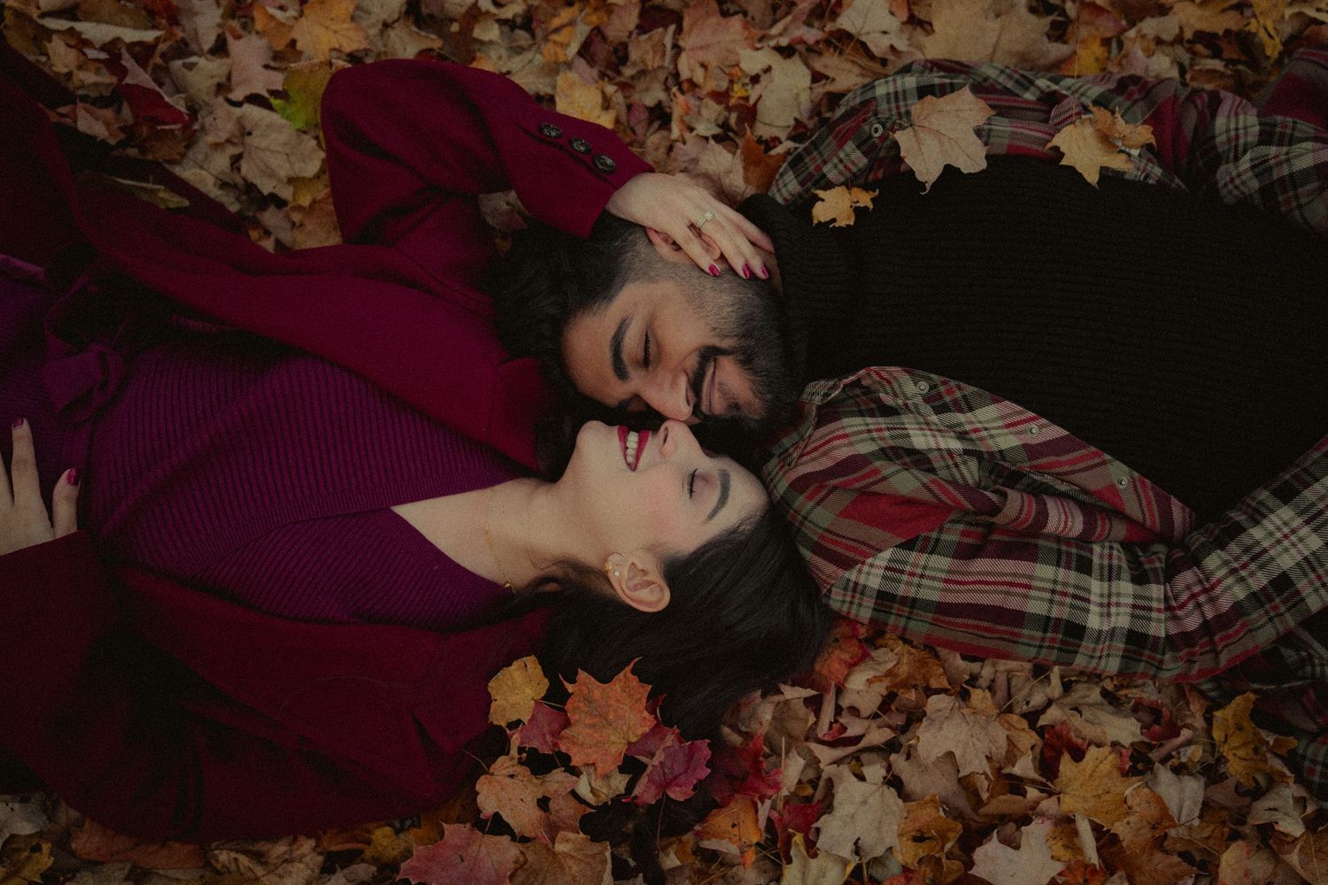 Romantic fall photography with couple lying among autumn leaves in burgundy attire.