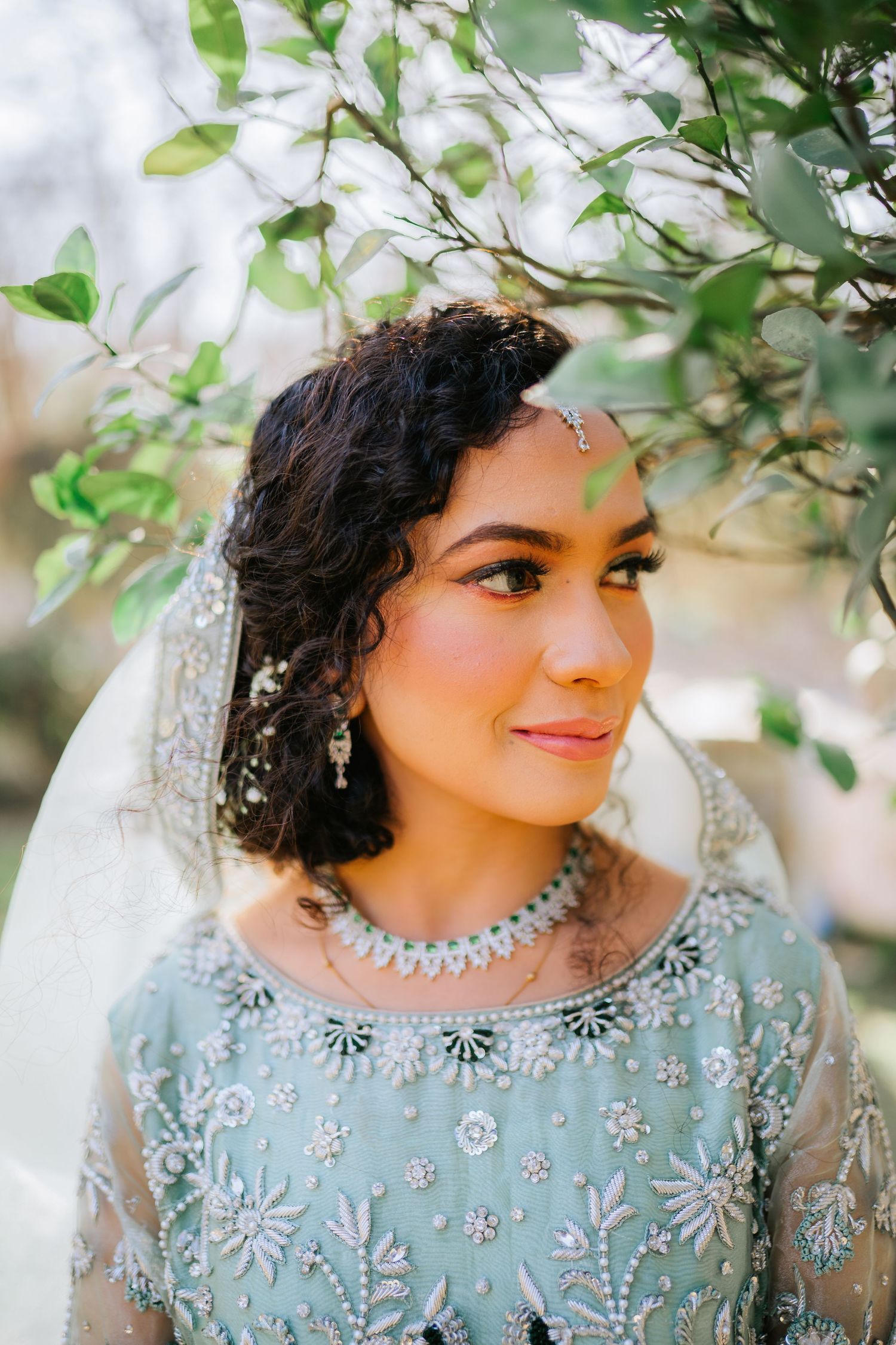 Bridal portrait featuring blue embellished dress and crystal jewelry against leafy background.