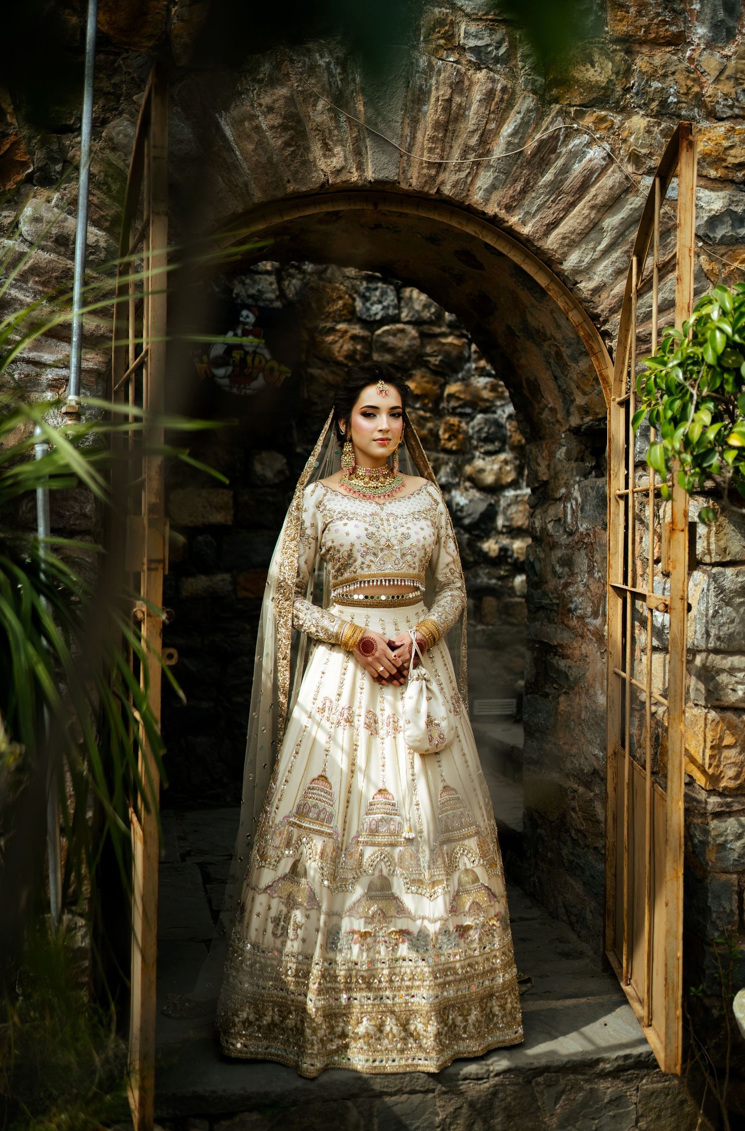 Ornate gold and cream bridal lehenga photographed in stone archway with dramatic lighting.
