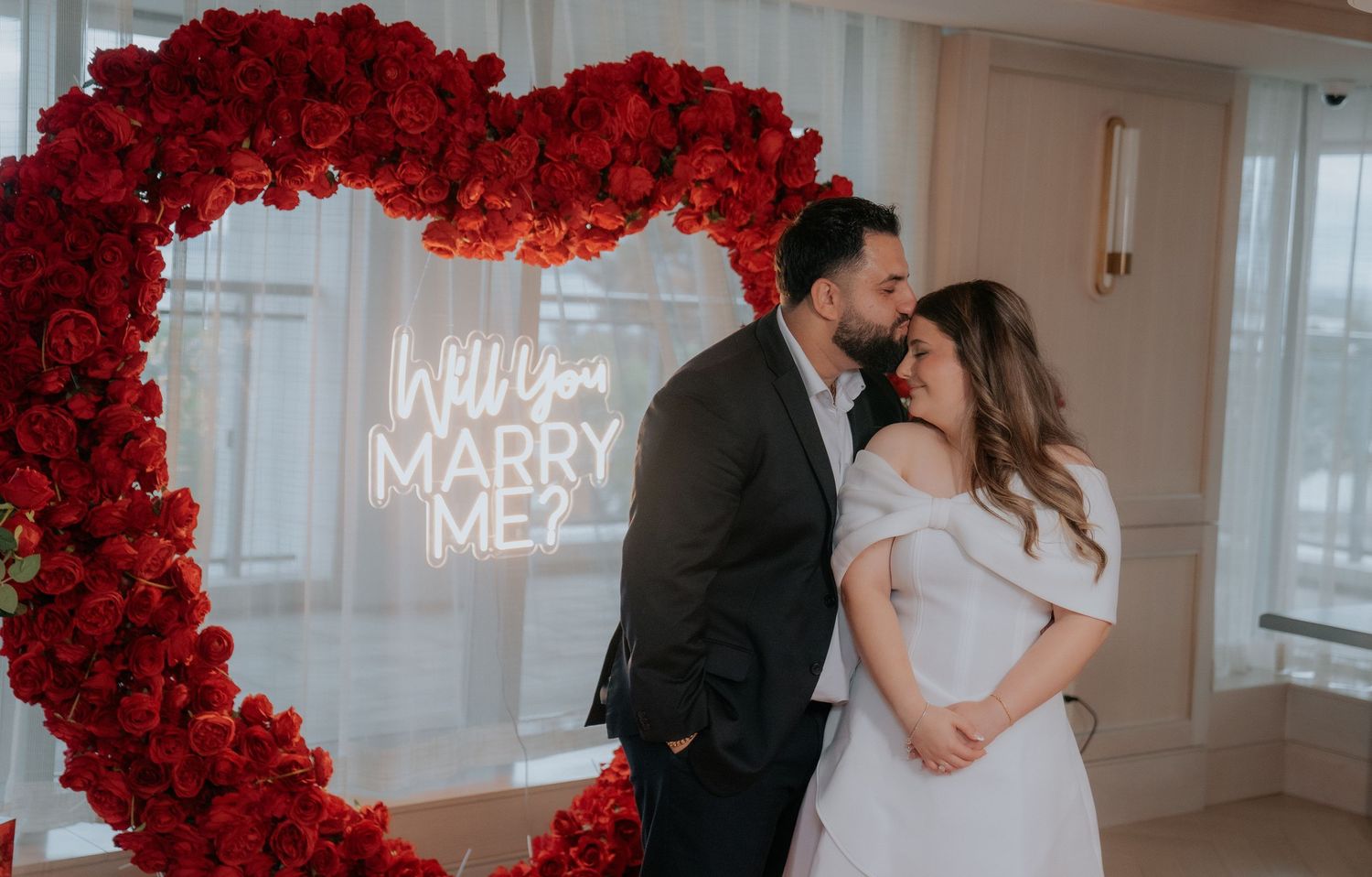 A romantic proposal scene under a dramatic arch of red roses with illuminated signage.