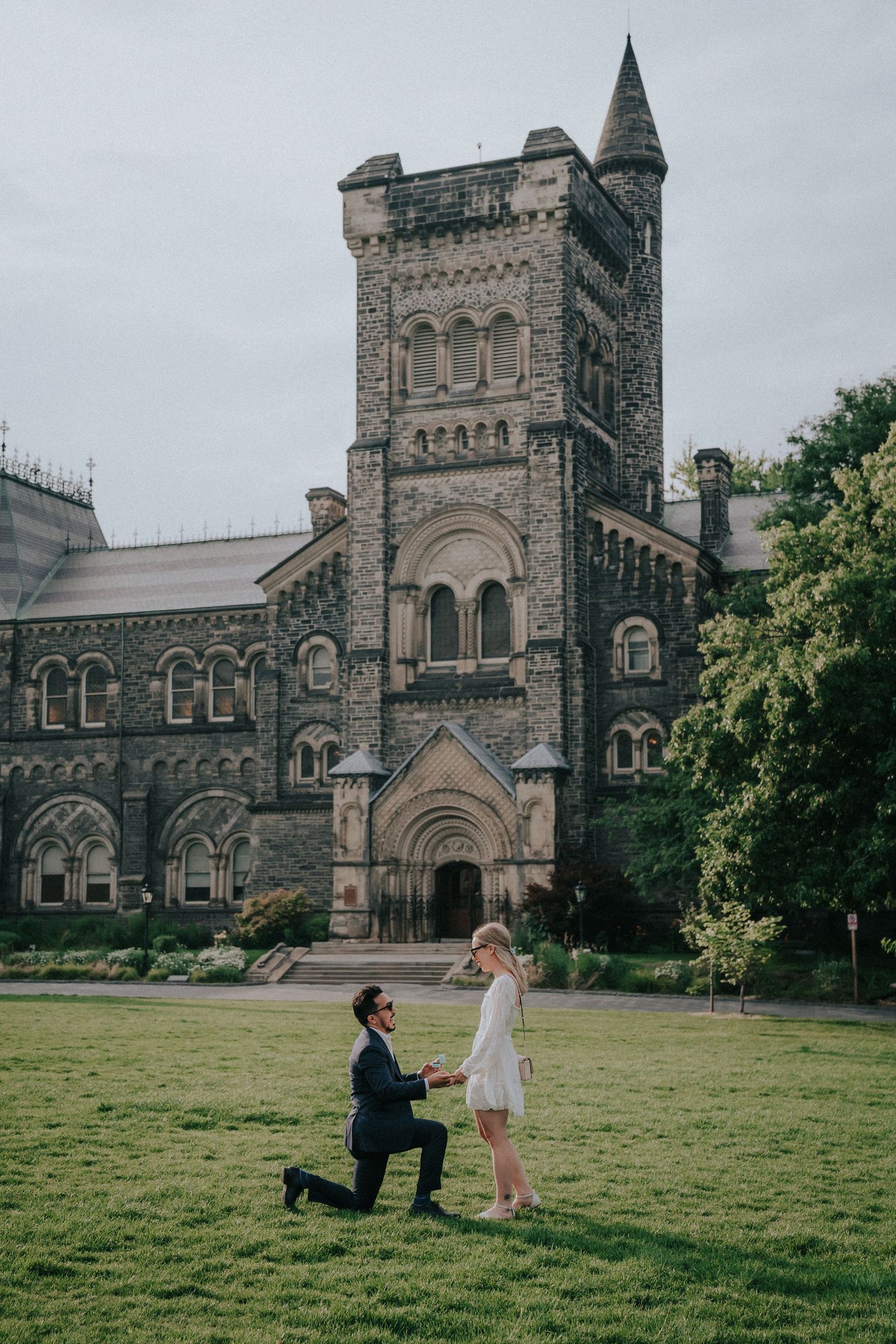 A proposal scene unfolds on the lawn in front of a grand Gothic university building.