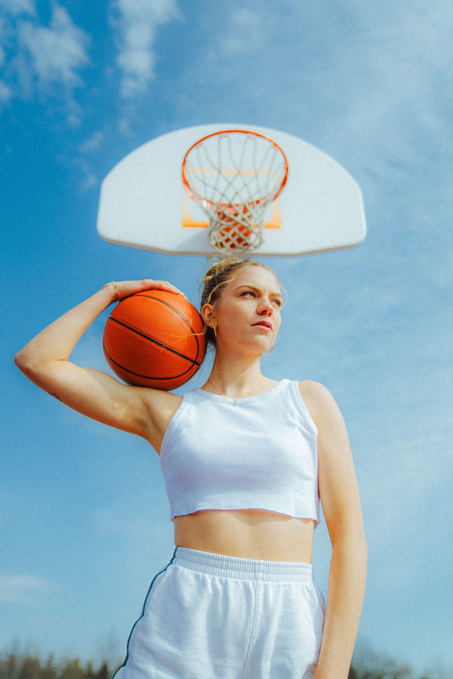 Athletic poses with basketball against blue sky and basketball hoop.