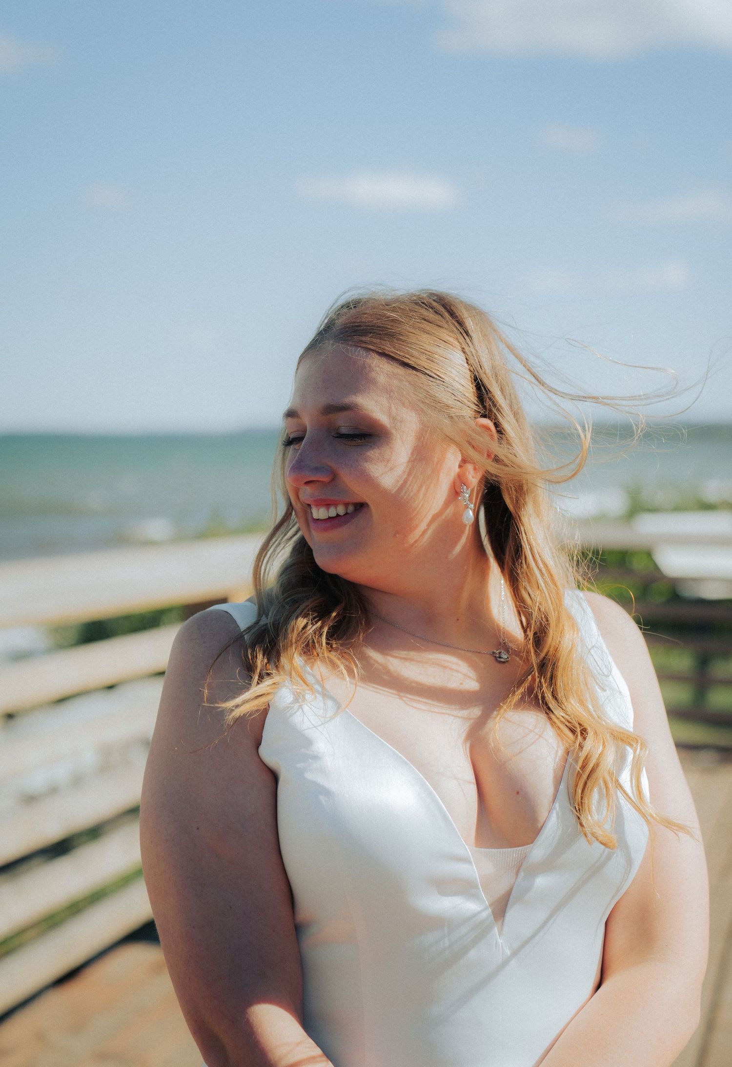 A bride in white dress laughs joyfully in bright sunlight by the ocean.